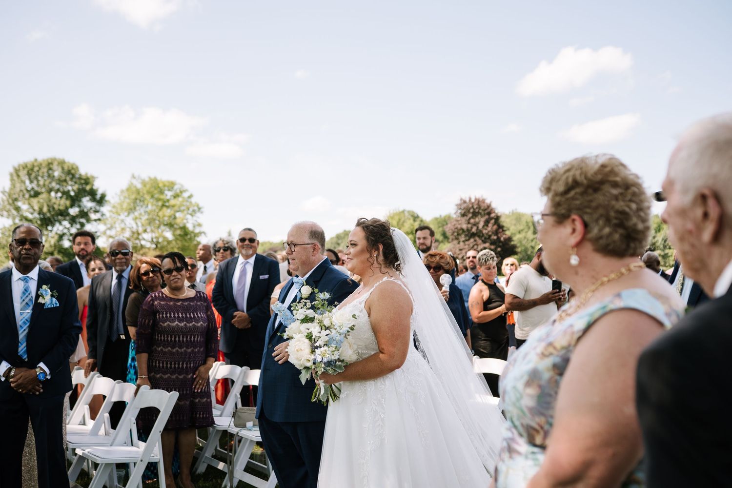 Wedding ceremony guests gather outdoors under trees with white chairs and floral decorations.