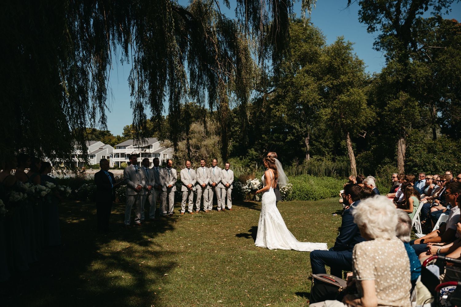 Outdoor wedding ceremony under a weeping willow tree with guests seated on white chairs.