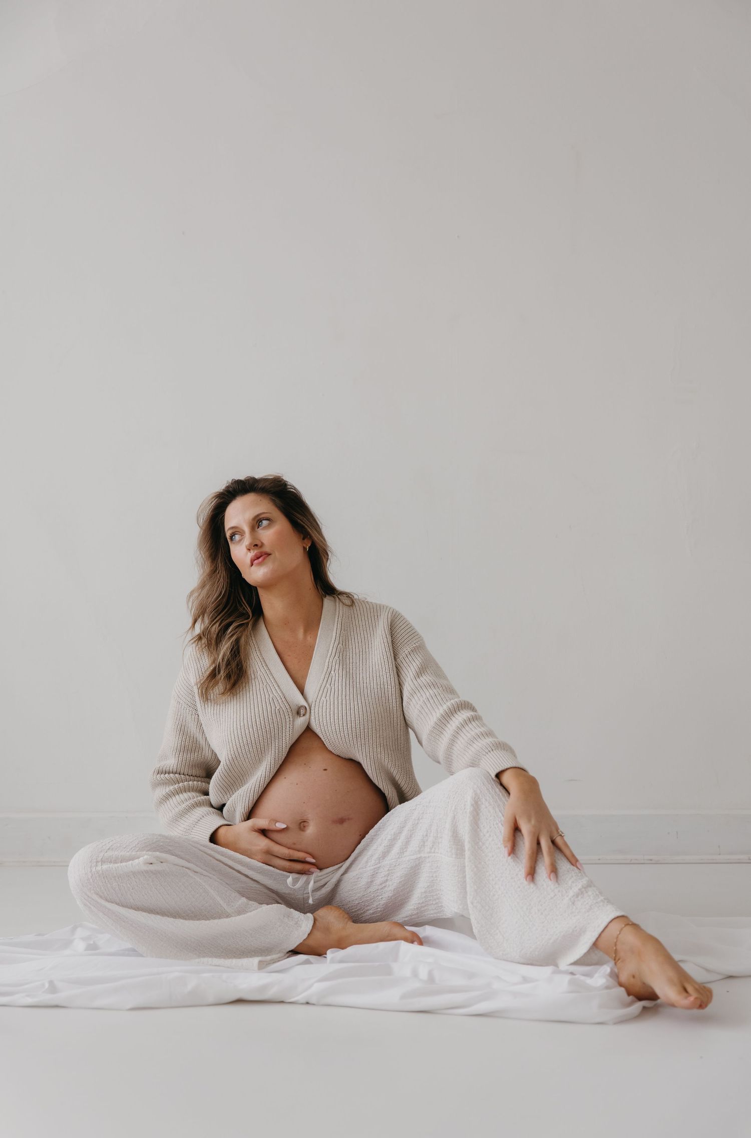Pregnant woman sitting on floor in beige cardigan and white pants, cradling bare bump, looking up serenely.