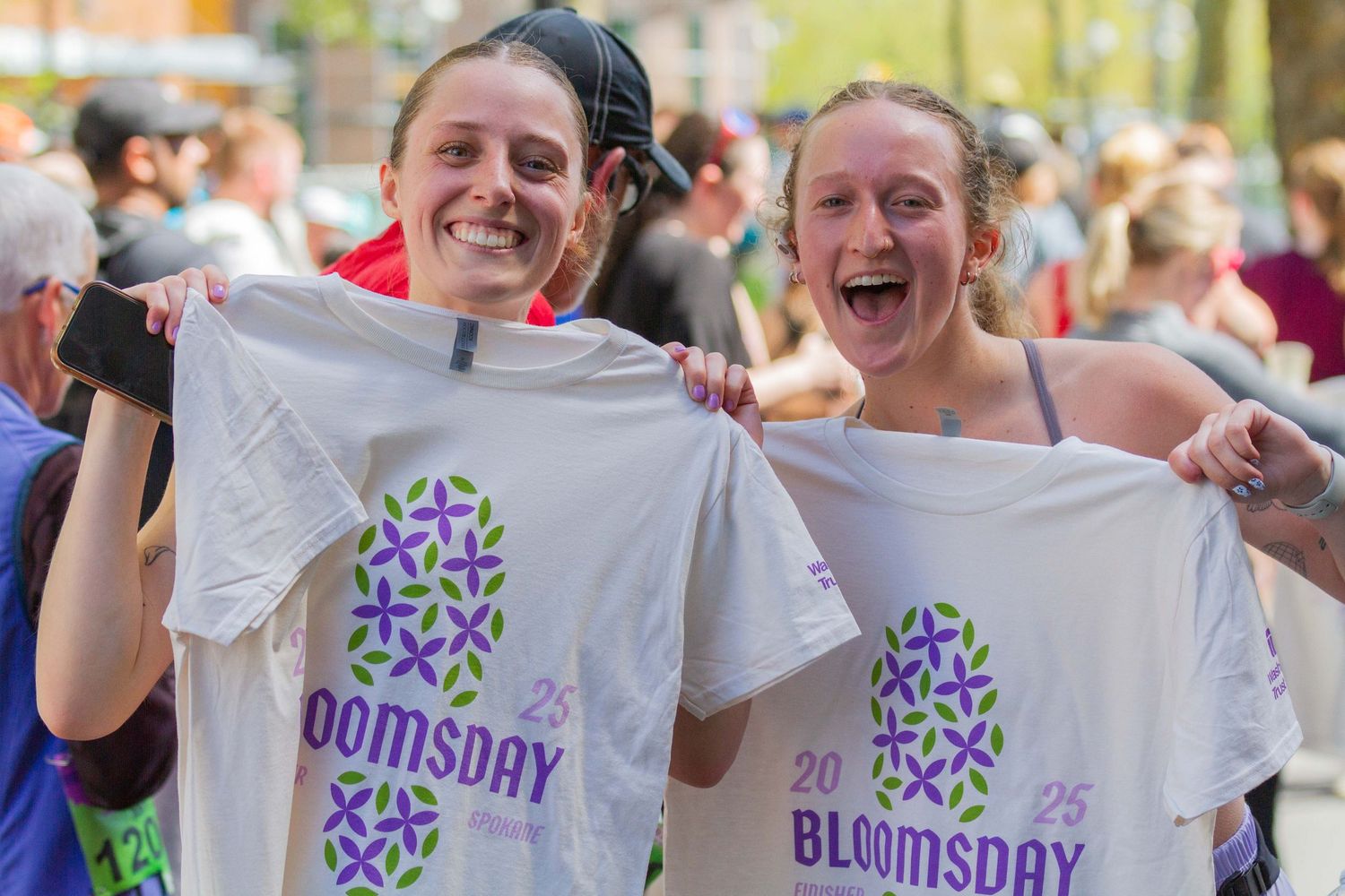 Two women smiling and holding up Bloomsday 25 race t-shirts outdoors after completing the event.