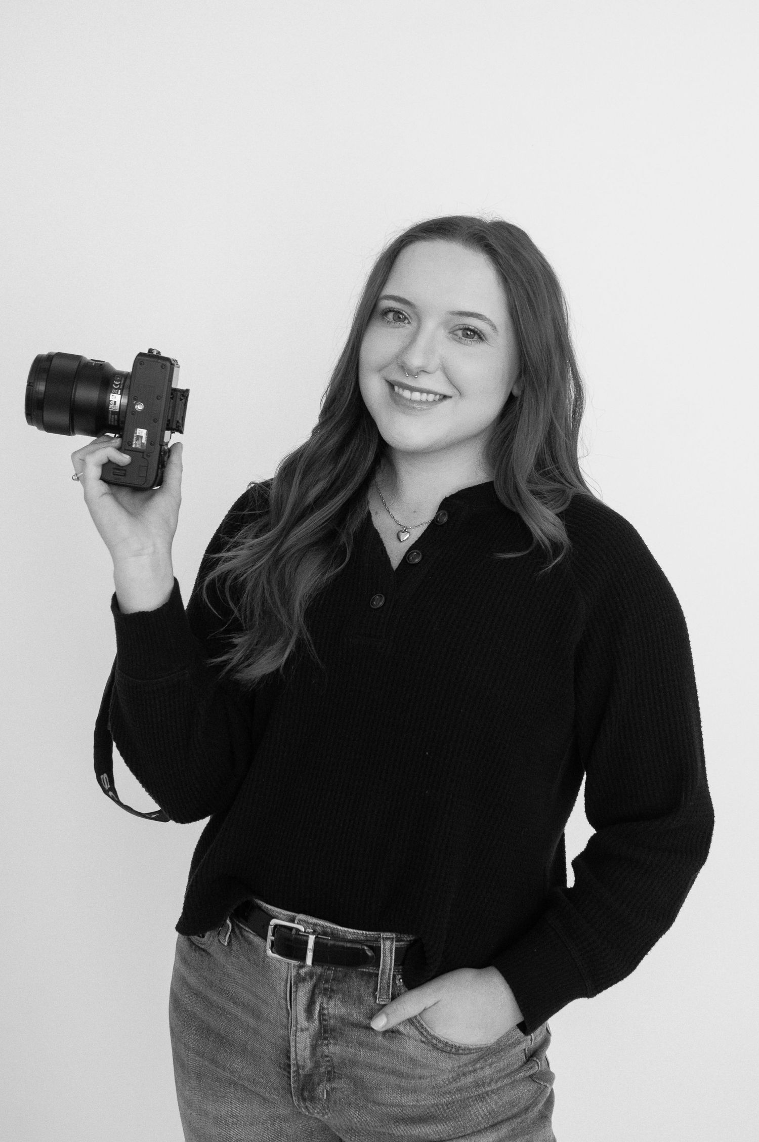 Black and white portrait of a smiling woman holding a camera, wearing a black top and jeans against a white background.
