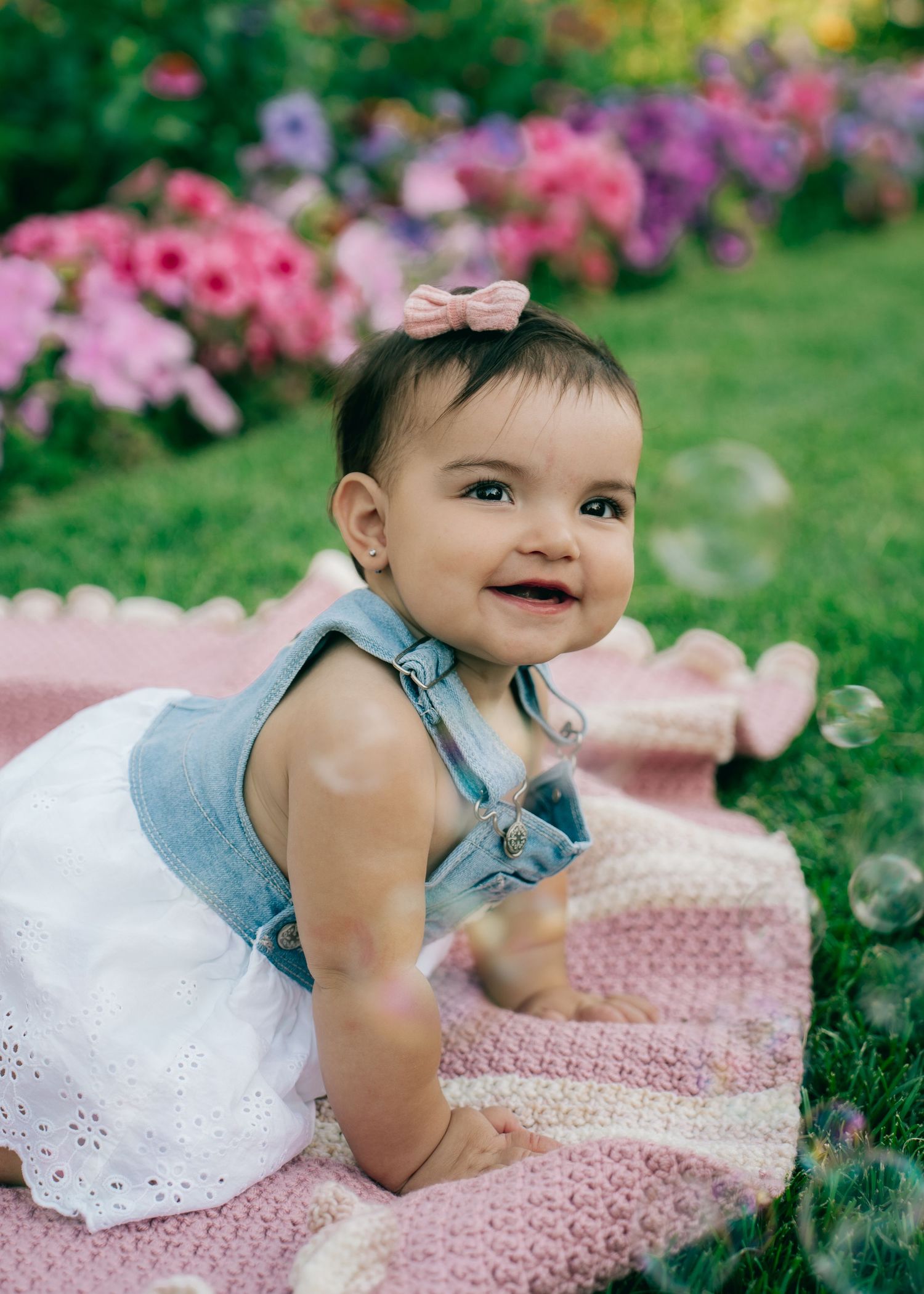 Baby in white dress and denim sitting on pink blanket with flowers in background.