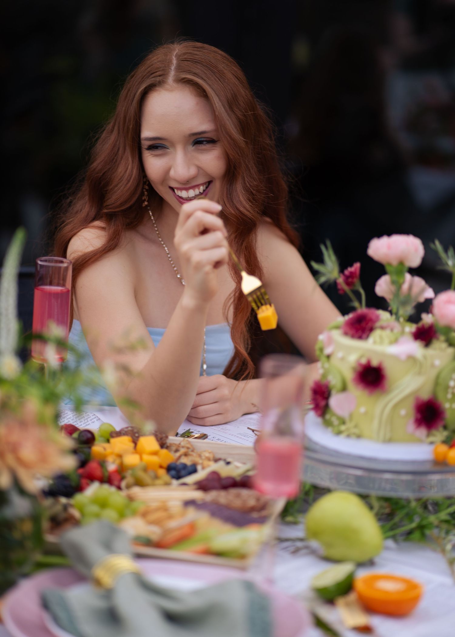 Smiling redhead woman eating at a floral outdoor table with colorful fruits, charcuterie, and a decorated cake.