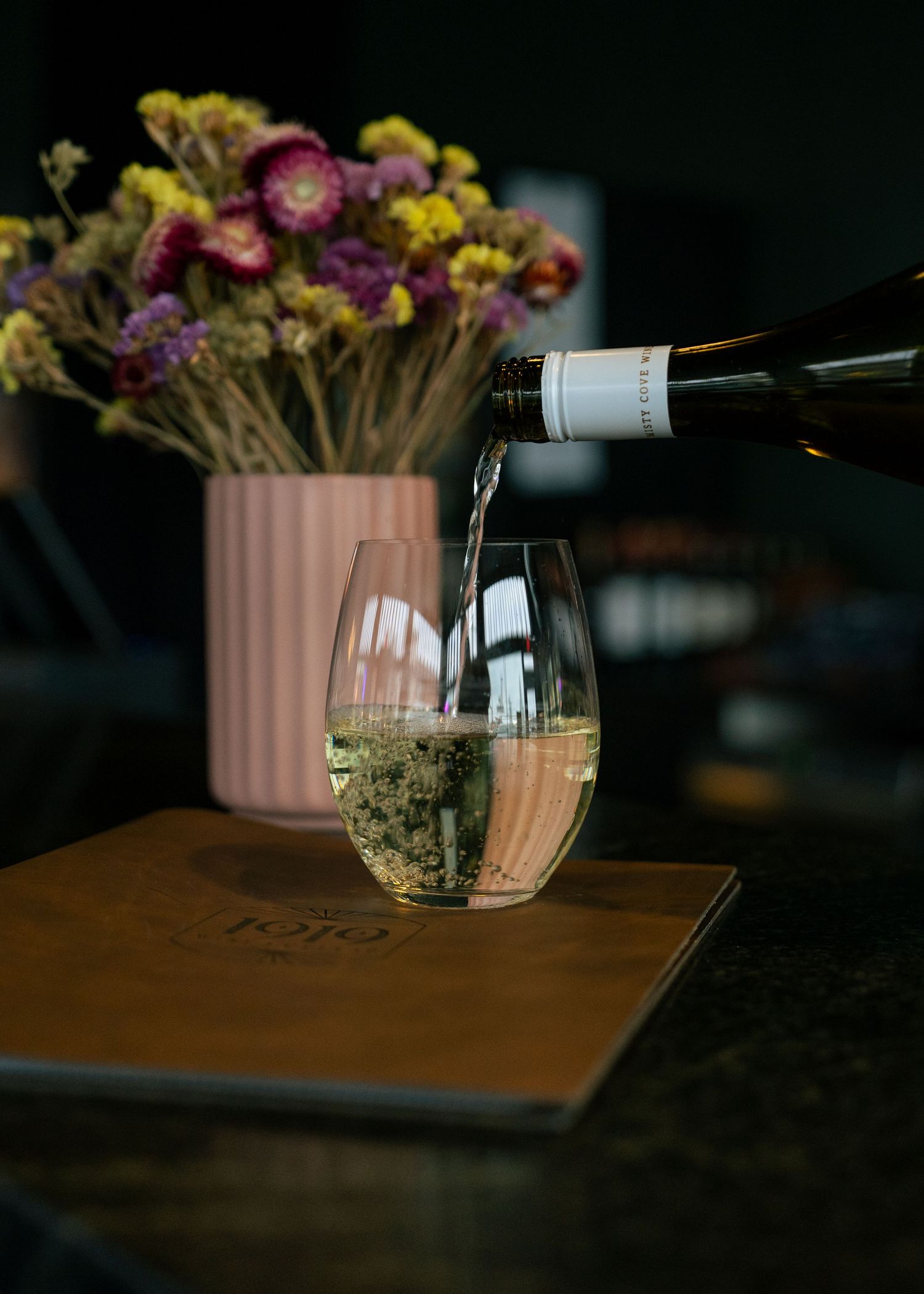Wine being poured into a glass with dried flowers in a pink vase in the background.