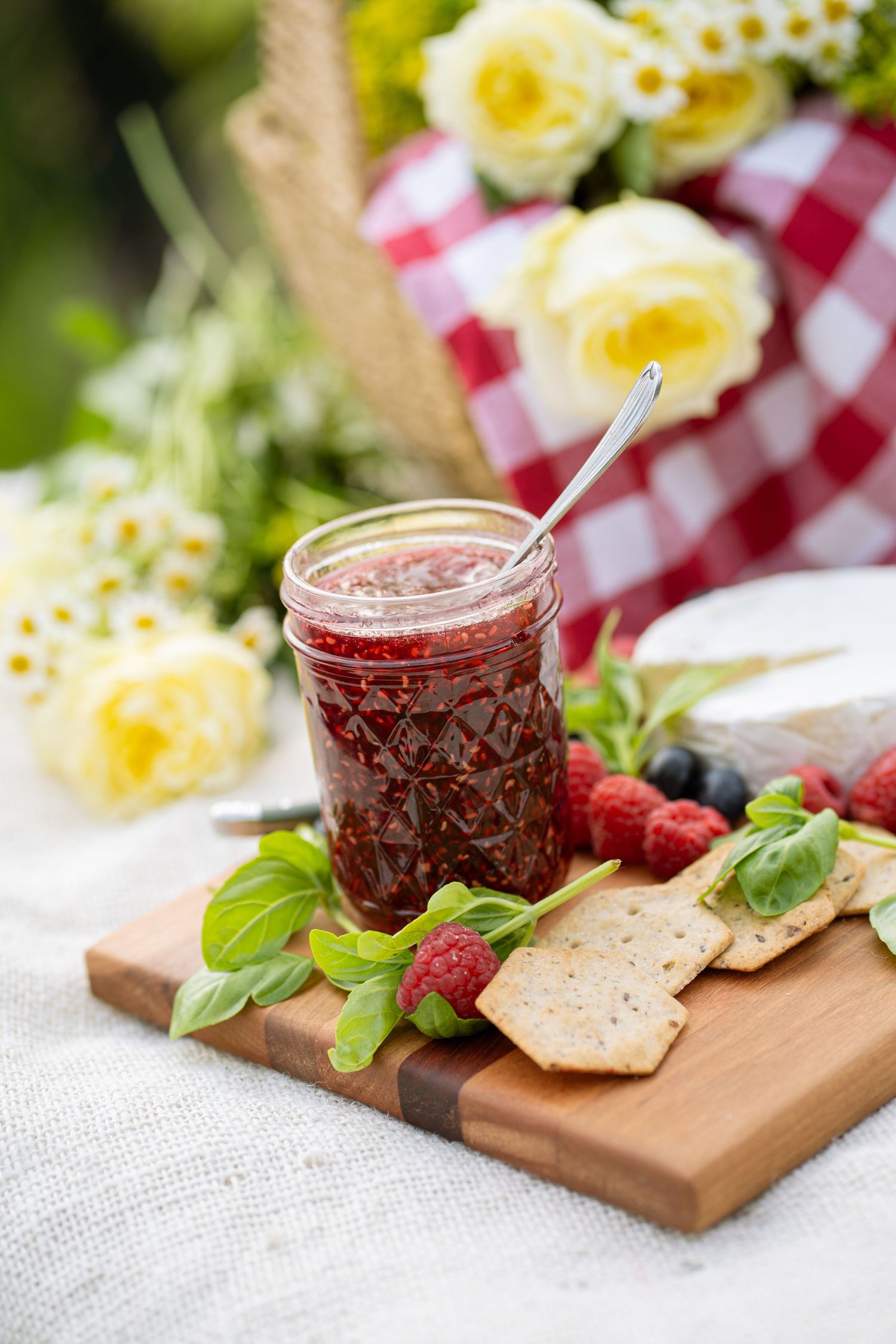 Raspberry jam jar with crackers, fresh berries, and cheese on a wooden board at an outdoor picnic with yellow roses.
