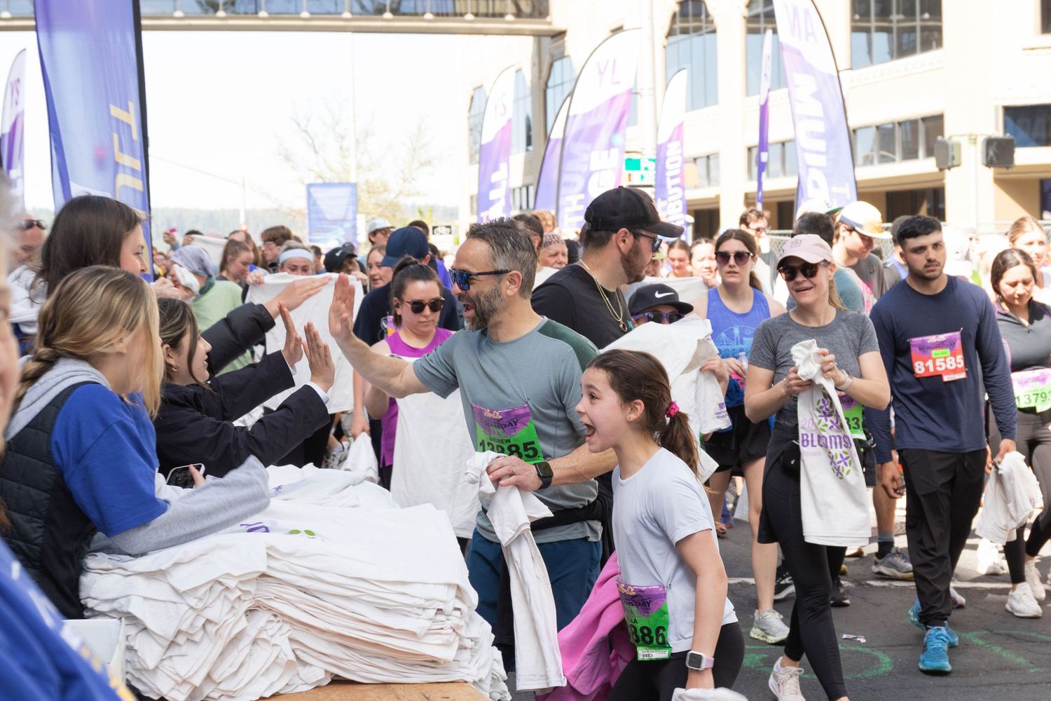Crowd of participants picking up white t-shirts at an outdoor charity run event with purple banners.