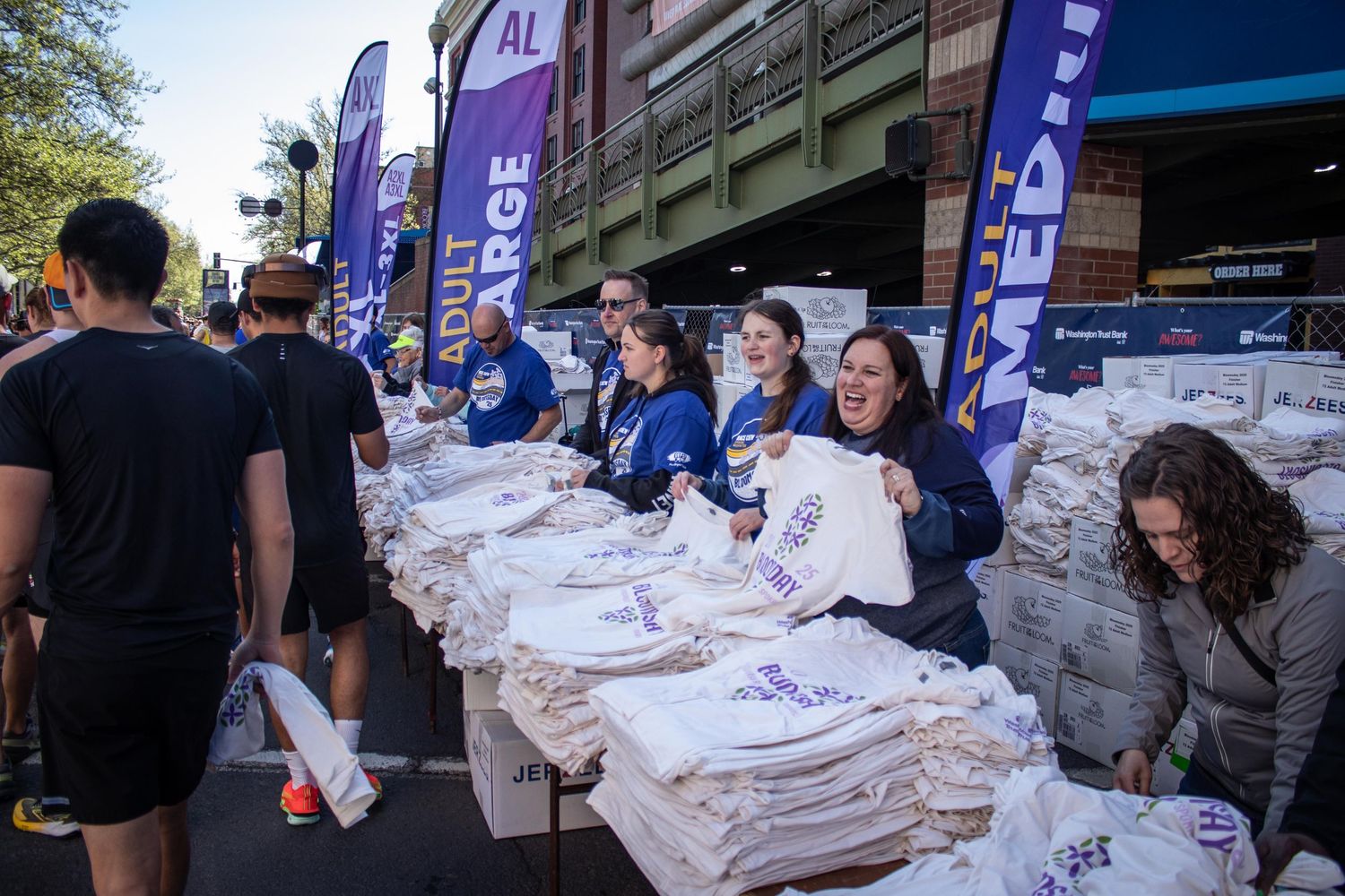 Volunteers in blue shirts distribute race t-shirts at an outdoor charity run event with purple banners.