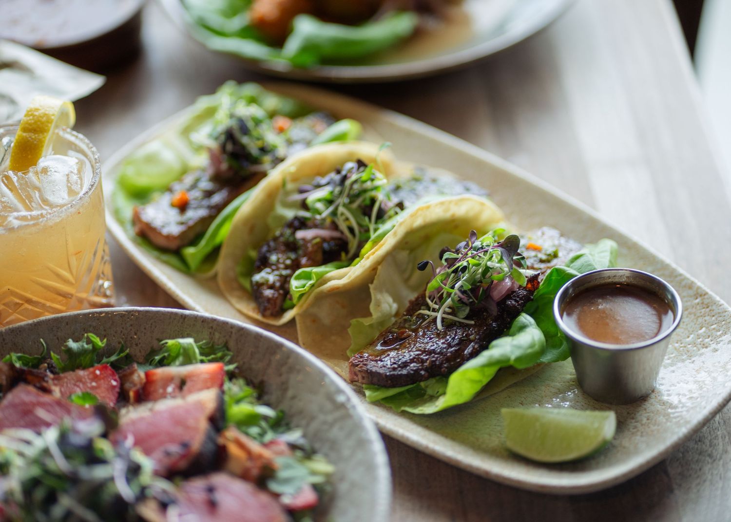 A plate of gourmet tacos with lettuce, meat, and sprouts served with dipping sauce and lime wedges on a wooden table.