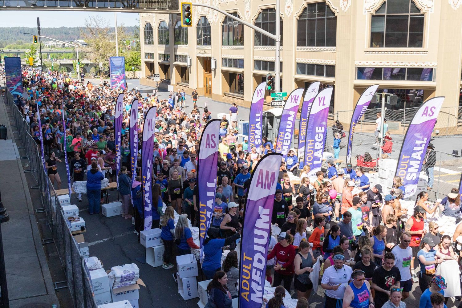 Large crowd of runners gather at a charity race start line with purple promotional banners on a sunny day.