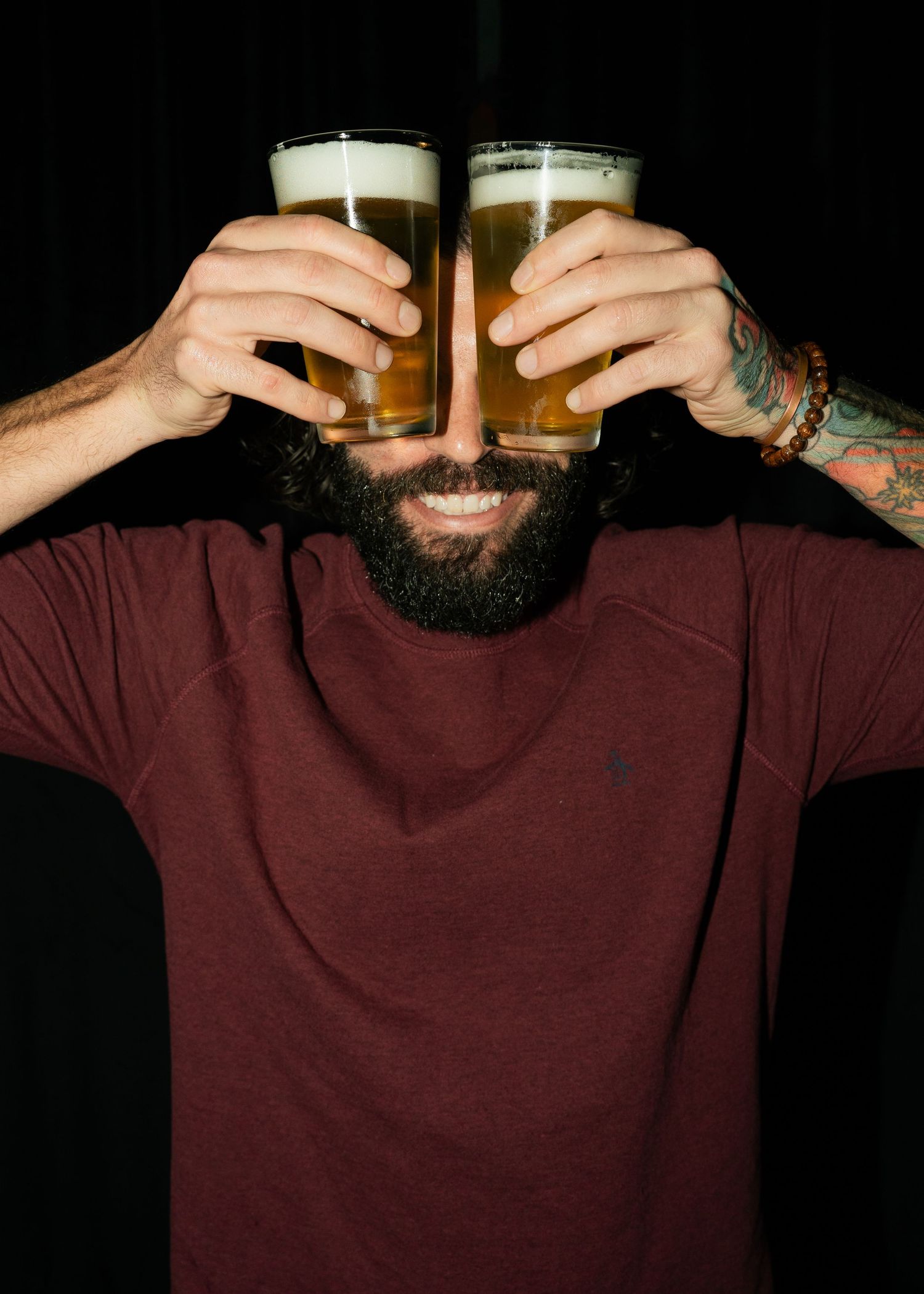 Person in burgundy shirt holding two glasses of beer against dark background.