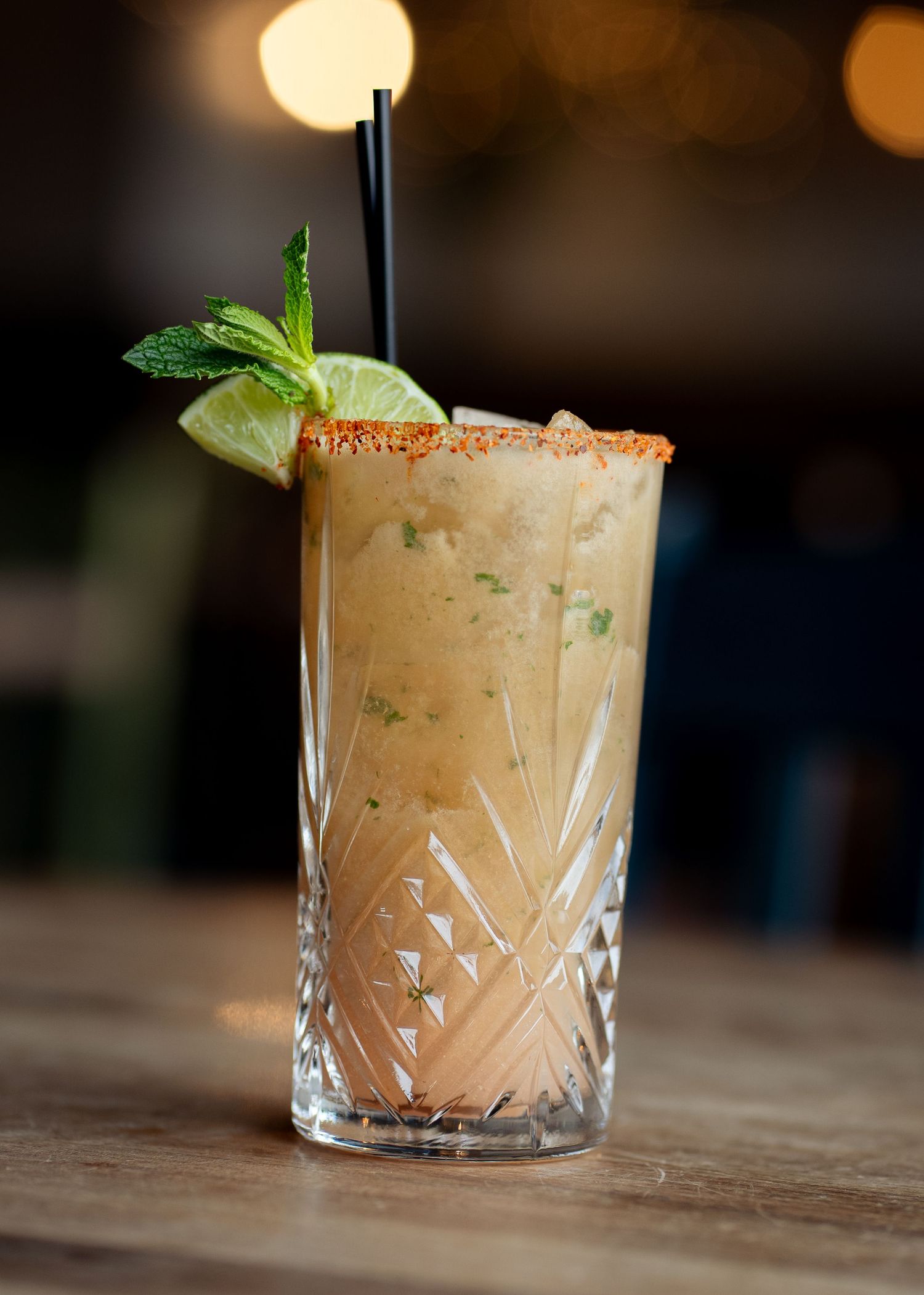 Spiced rim cocktail in crystal highball glass with lime wedge, mint garnish, and black straw on wooden bar.