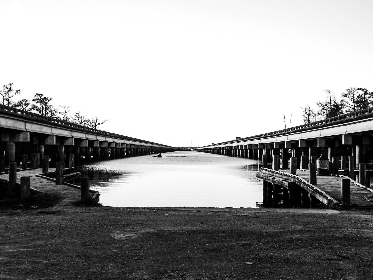 Black and white photo of a symmetrical twin bridge over calm water with concrete pillars and bare trees in the background.
