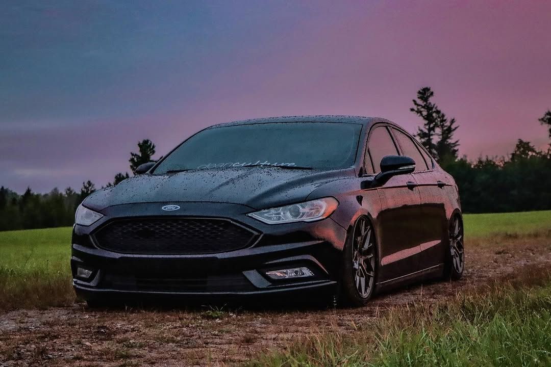 Black Ford Fusion SE sedan parked on dirt road at dusk with dramatic pink and purple sky backdrop.