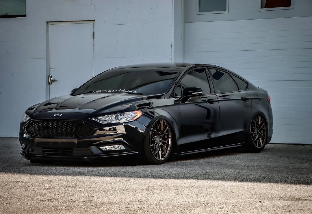 Black Ford Fusion sport sedan with custom lowered stance and dark bronze wheels parked outside a garage.