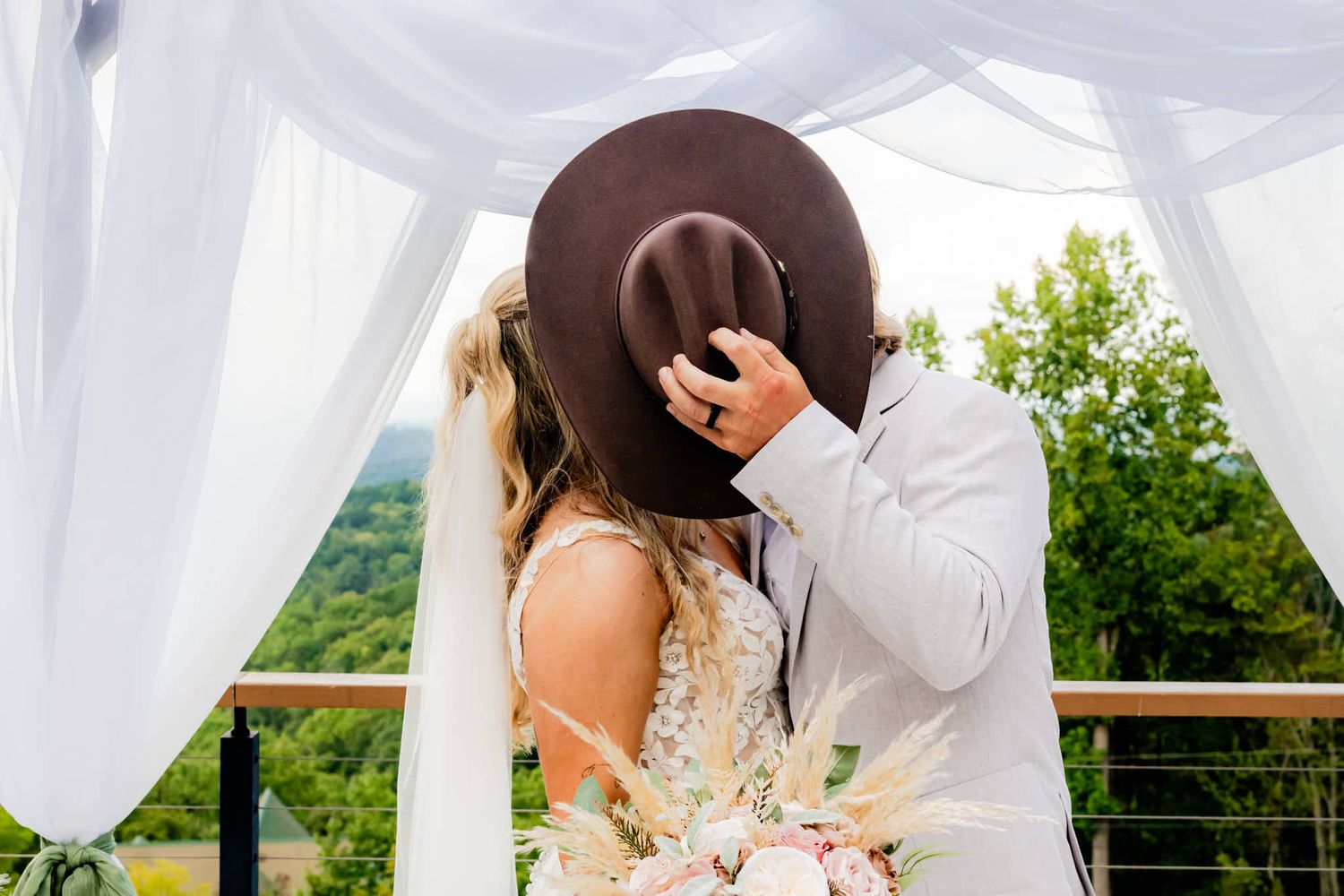 Bride and groom sharing a kiss hidden behind a brown wide-brim hat at an outdoor wedding ceremony.