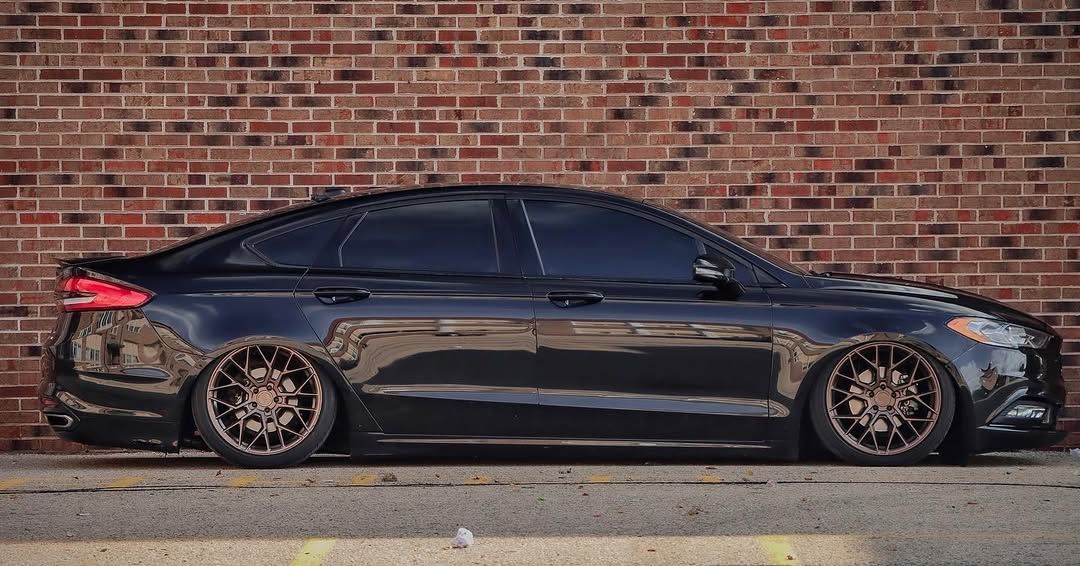 Lowered black Ford Fusion with bronze wheels parked against a red brick wall, showcasing a custom stance modification.