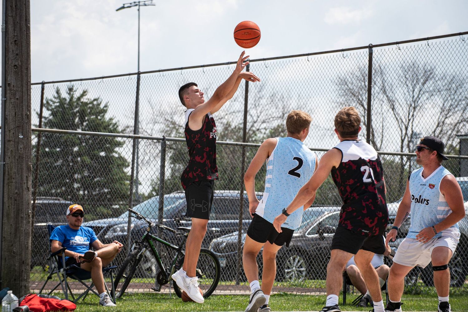 A player in dark jersey shoots a basketball while opponents in light uniforms defend on an outdoor court.