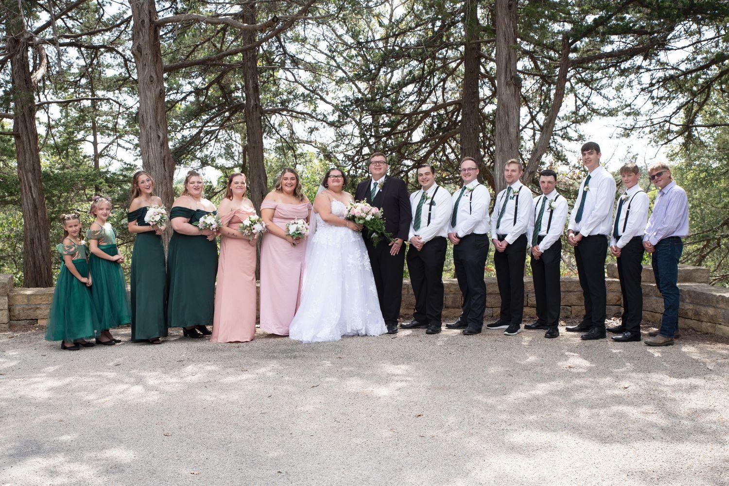 Wedding party poses under pine trees with bridesmaids in pink and green gowns and groomsmen in black in Lanesboro MN.