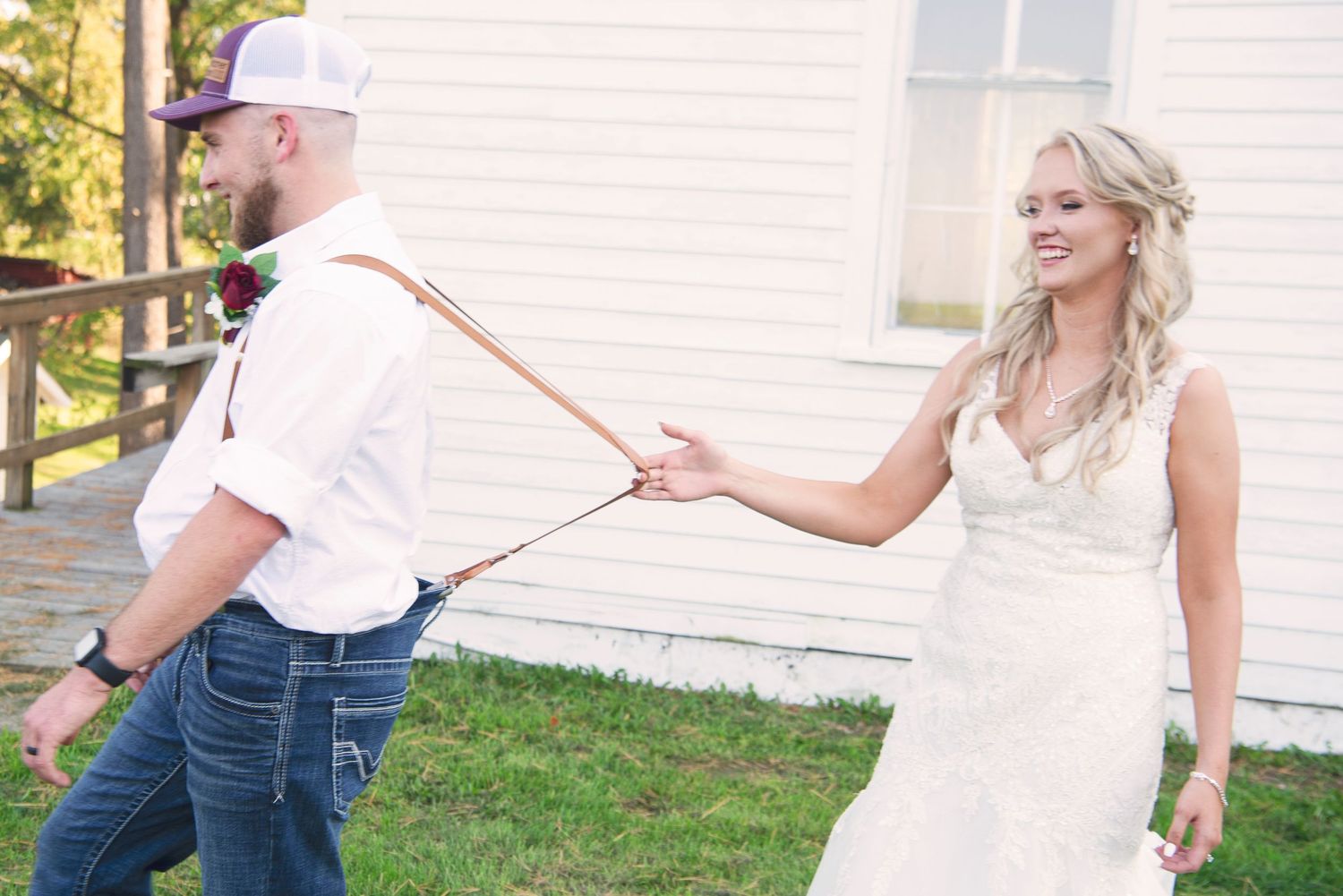 Smiling bride in white lace dress playfully pulls groom by suspenders outdoors at country wedding.