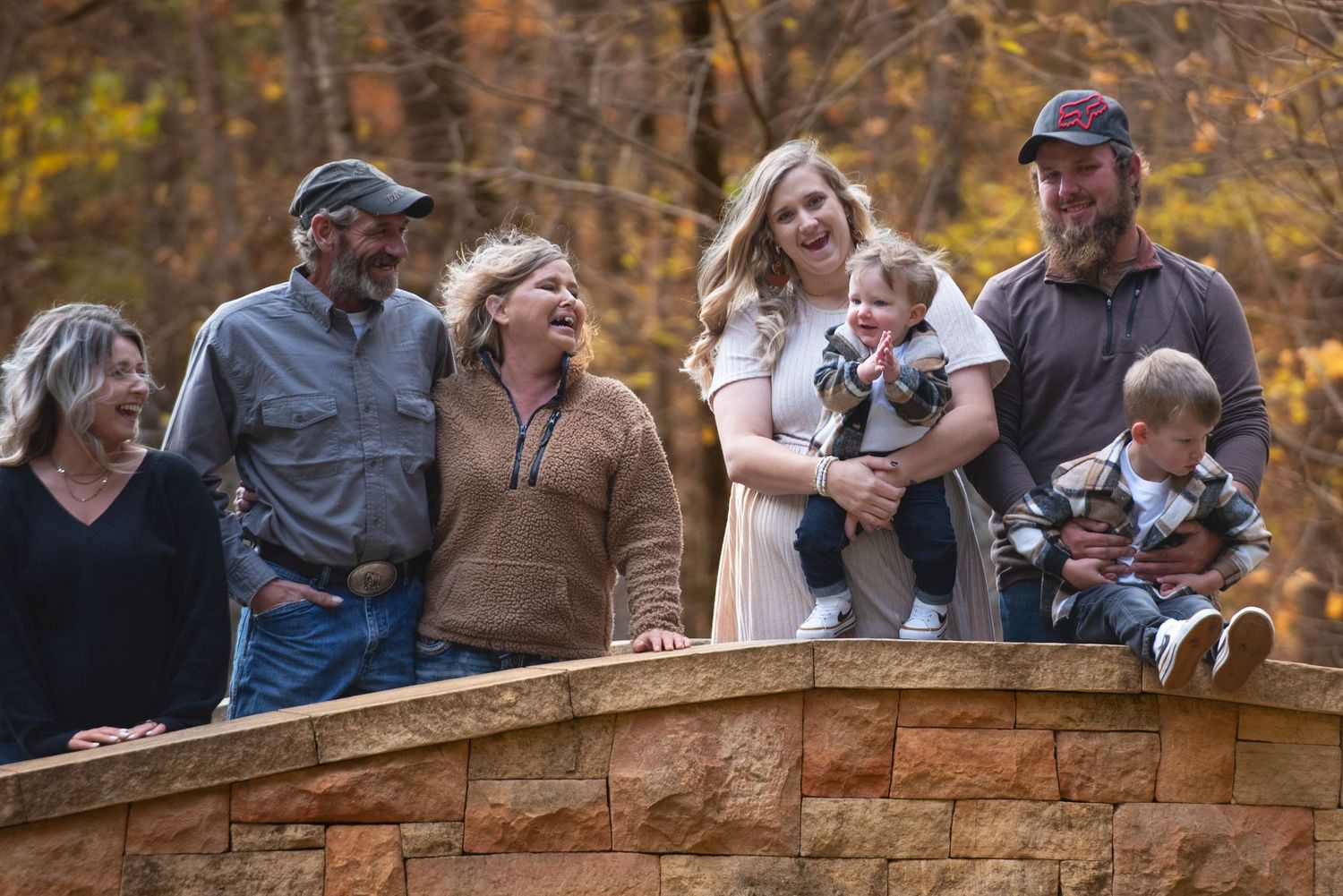 Multigenerational family laughing together on a stone bridge wall amid golden fall trees in outdoor setting.