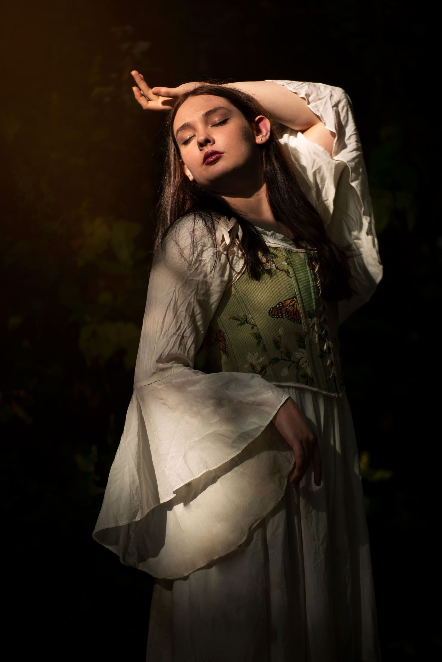 Young woman in flowing white and green medieval dress posing dramatically in dark forest.