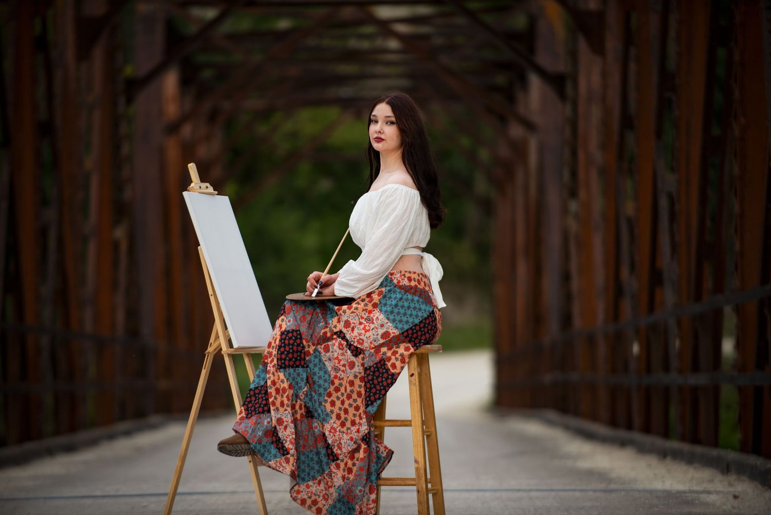 Elegant brunette woman in bohemian skirt sitting at an easel painting on a rustic bridge.