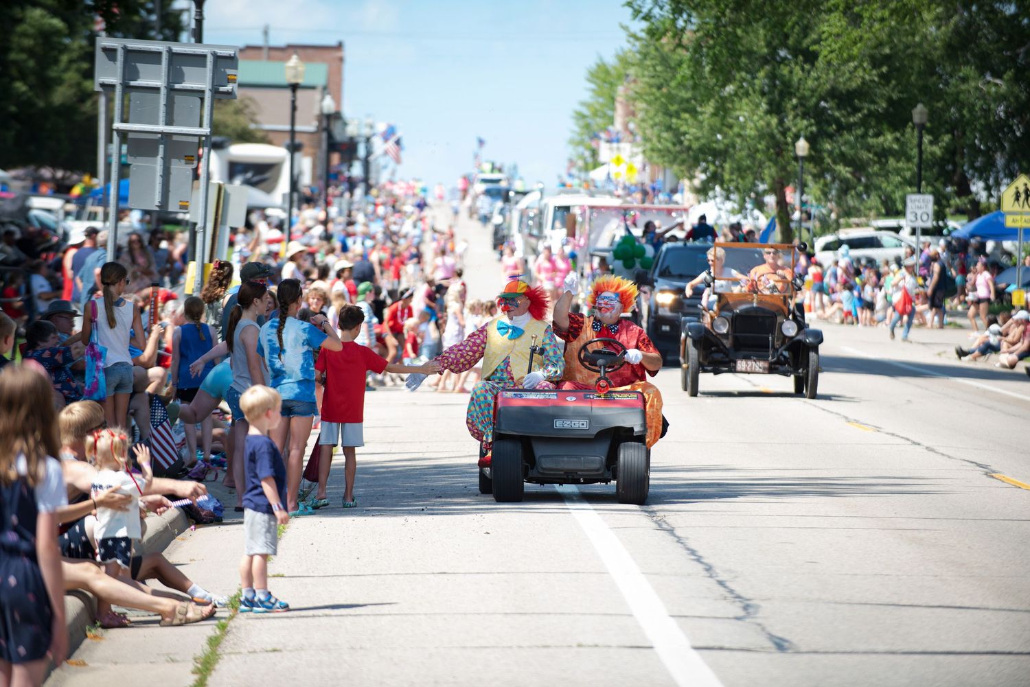 A colorful clown drives a small vehicle past cheering crowds lining the street at a Fourth of July parade at Harmony MN.