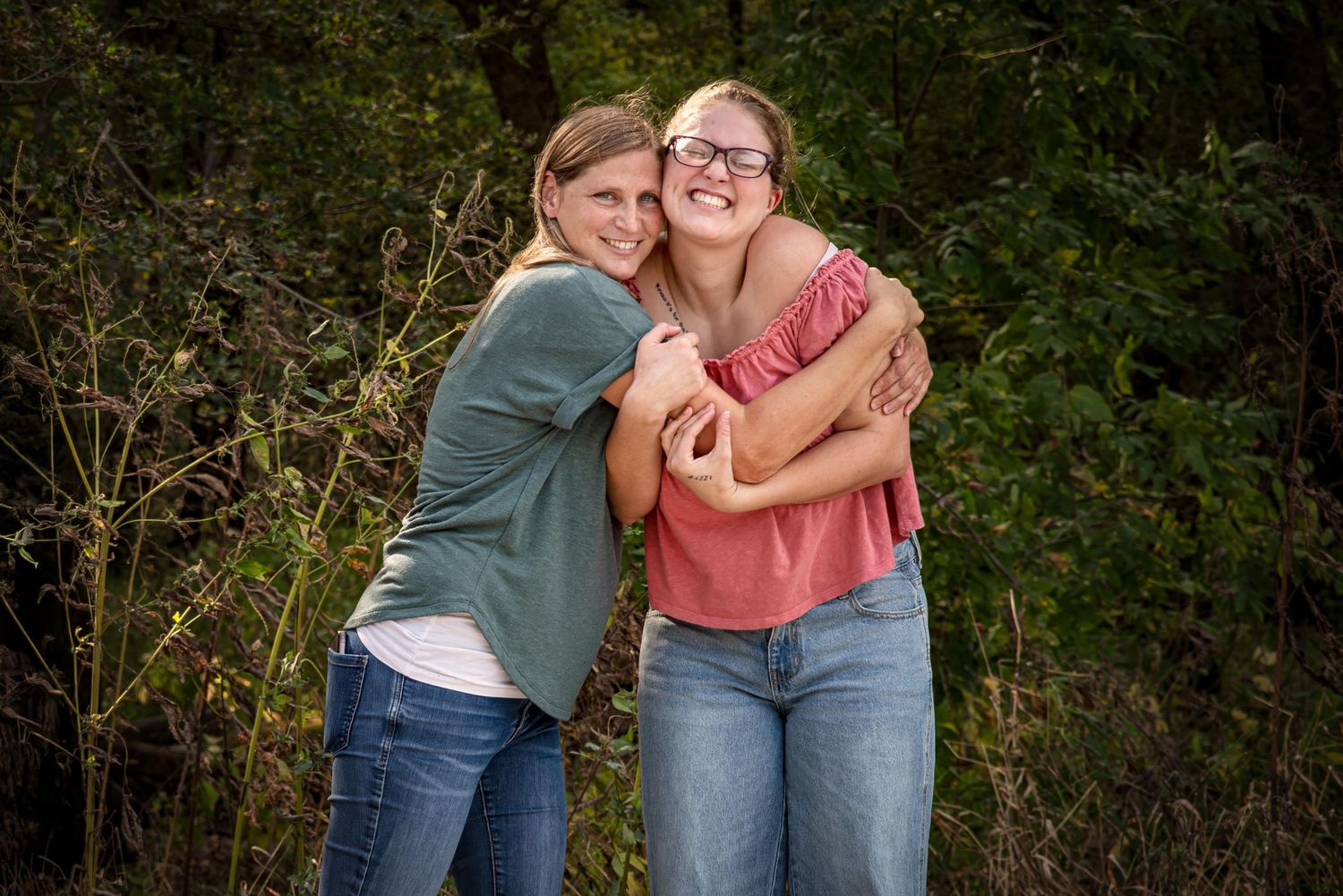 Two smiling teenage girls share a warm hug outdoors surrounded by green foliage.