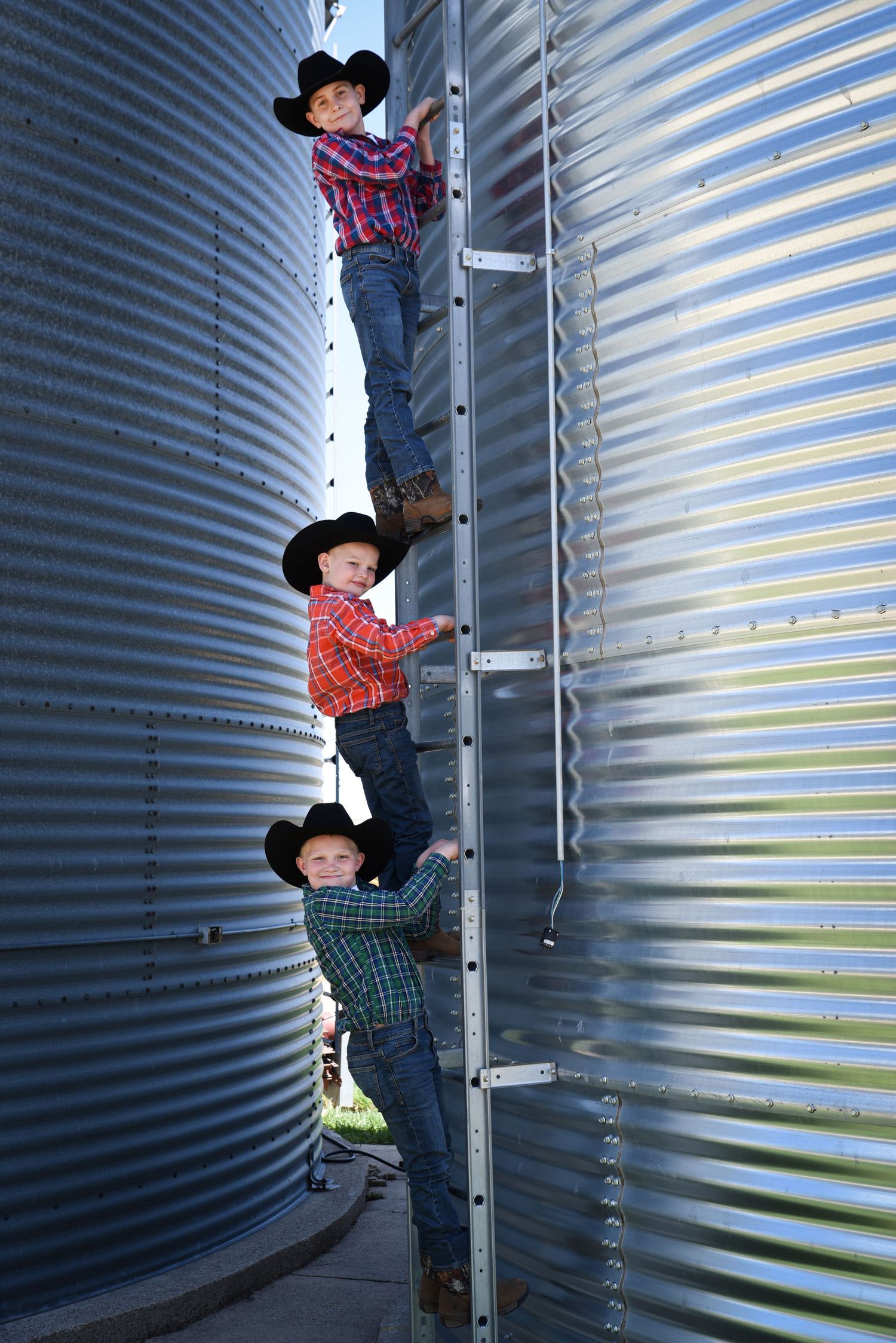 Three boys wearing cowboy hats and plaid shirts climbing a metal grain silo ladder on a farm.