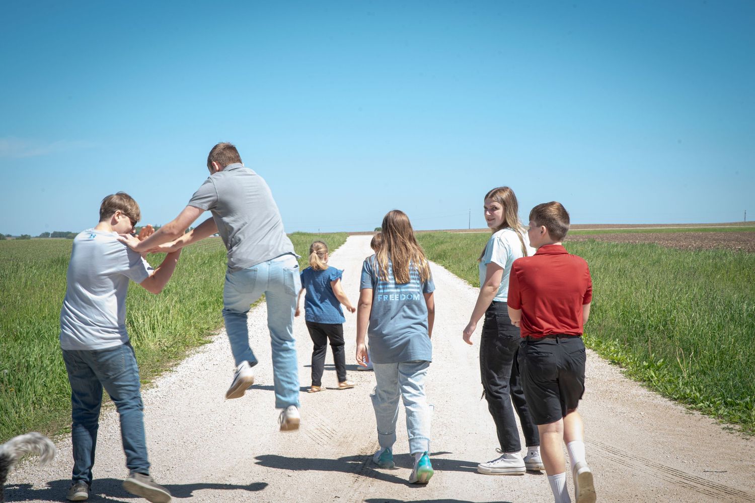 Group of children walking down a rural gravel road surrounded by green fields.