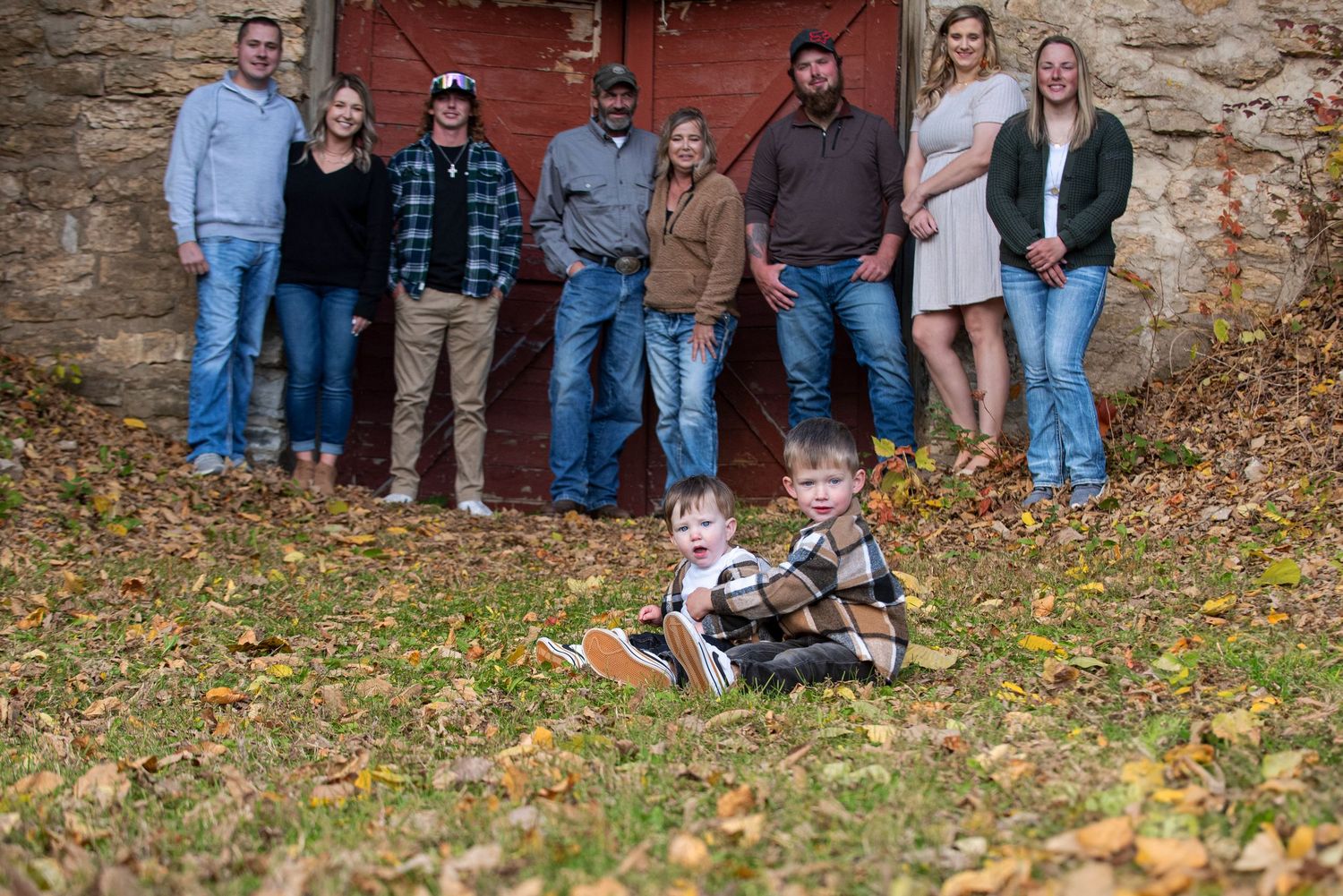 Extended family portrait with two young children sitting in fallen leaves before a rustic red barn door.