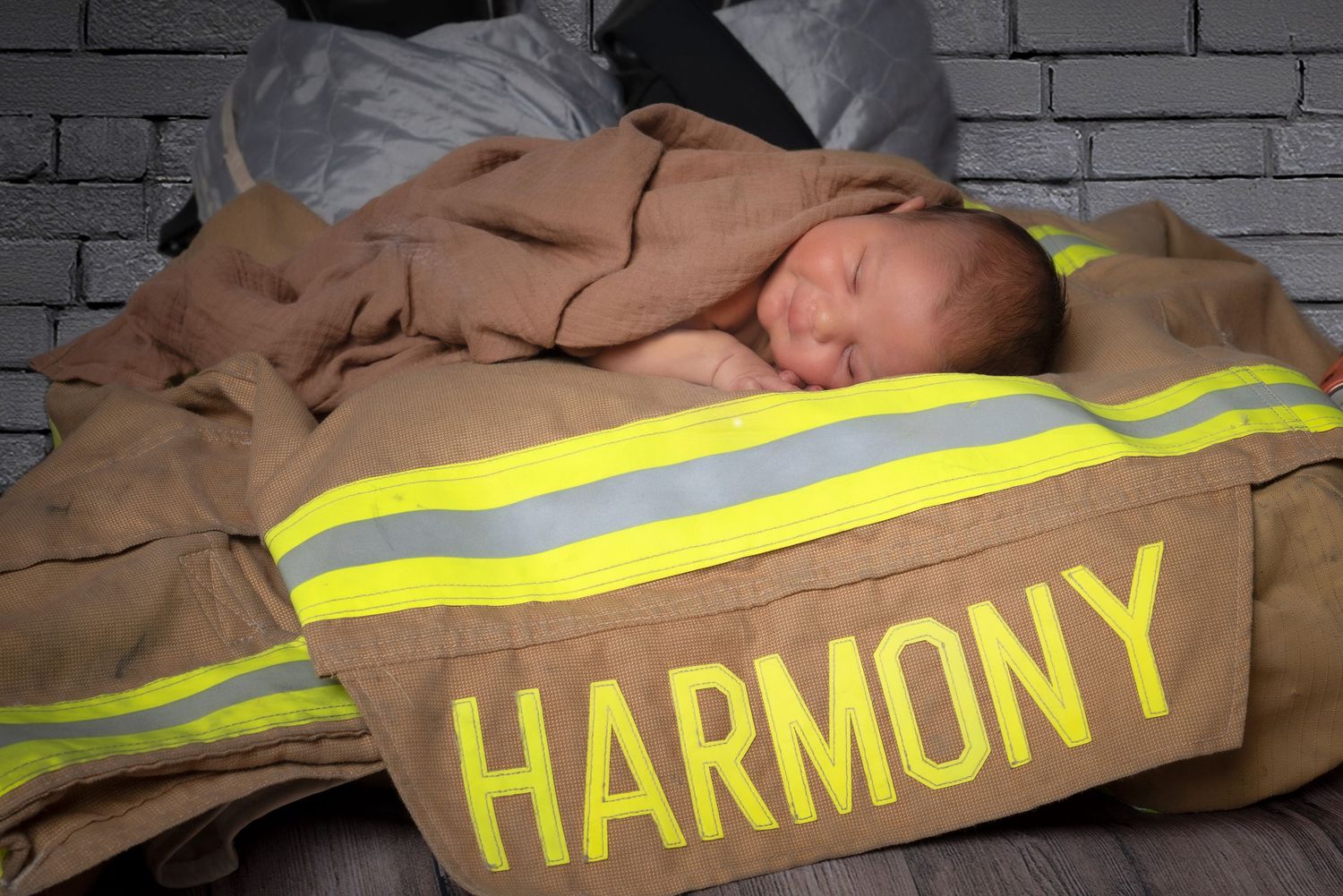 Newborn baby sleeping peacefully wrapped in a firefighter jacket with the name Harmony printed in yellow letters.