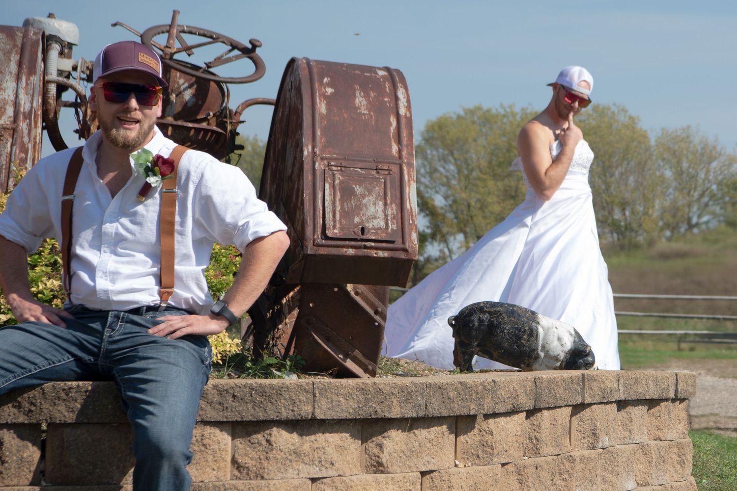 Bride and groom posing casually near rustic farm equipment during outdoor wedding celebration.