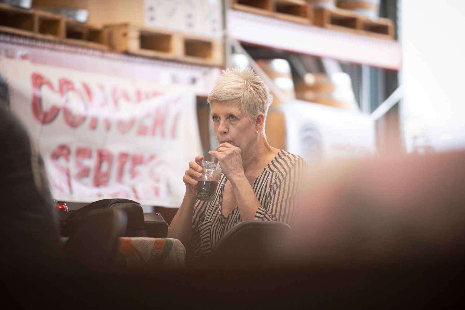 Woman with short blonde hair sitting and drinking a glass of water in front of a Concert Series banner at Harmony Spirits..