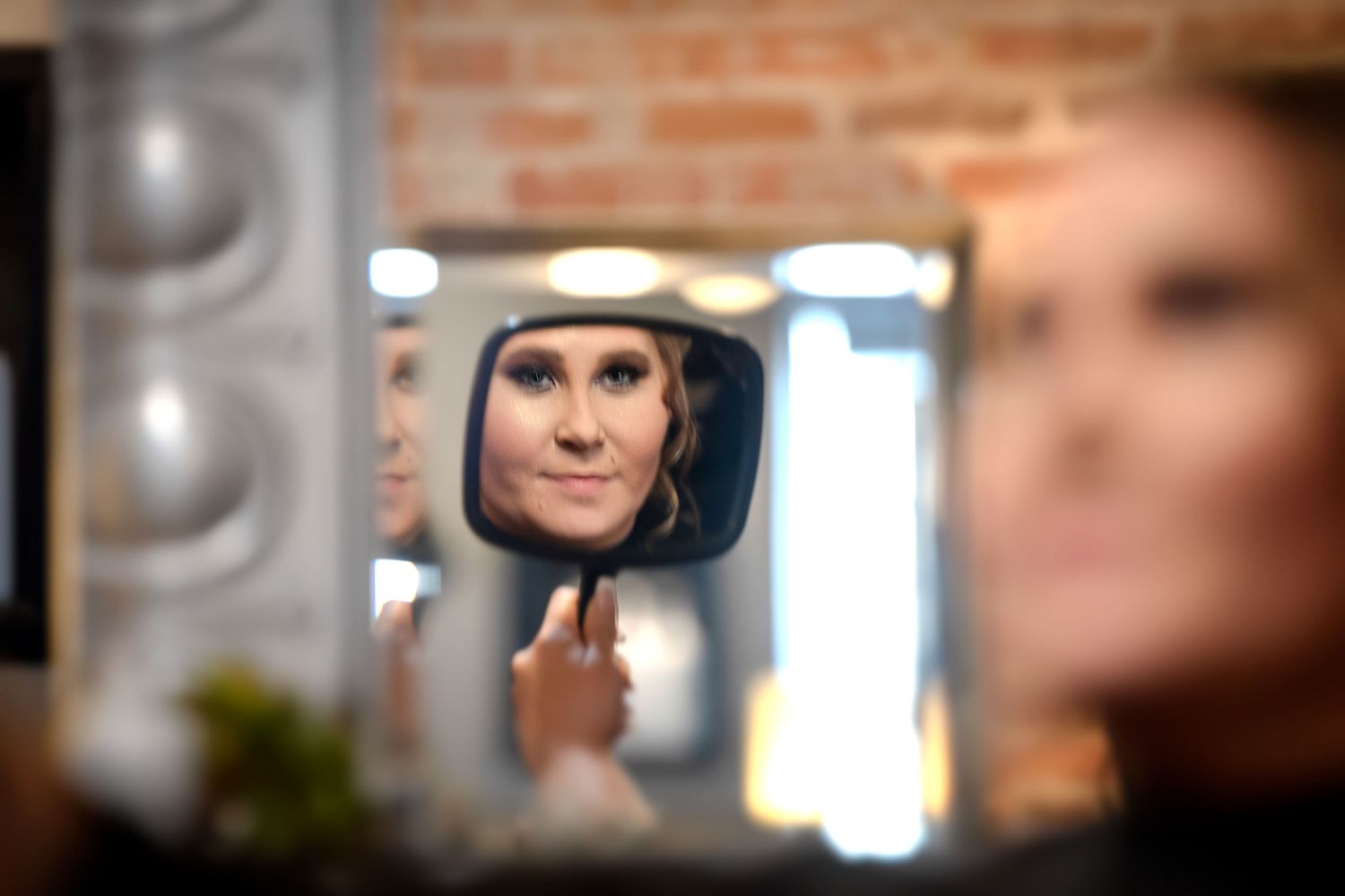 A woman examines her makeup in a handheld mirror at a vanity with studio lights.