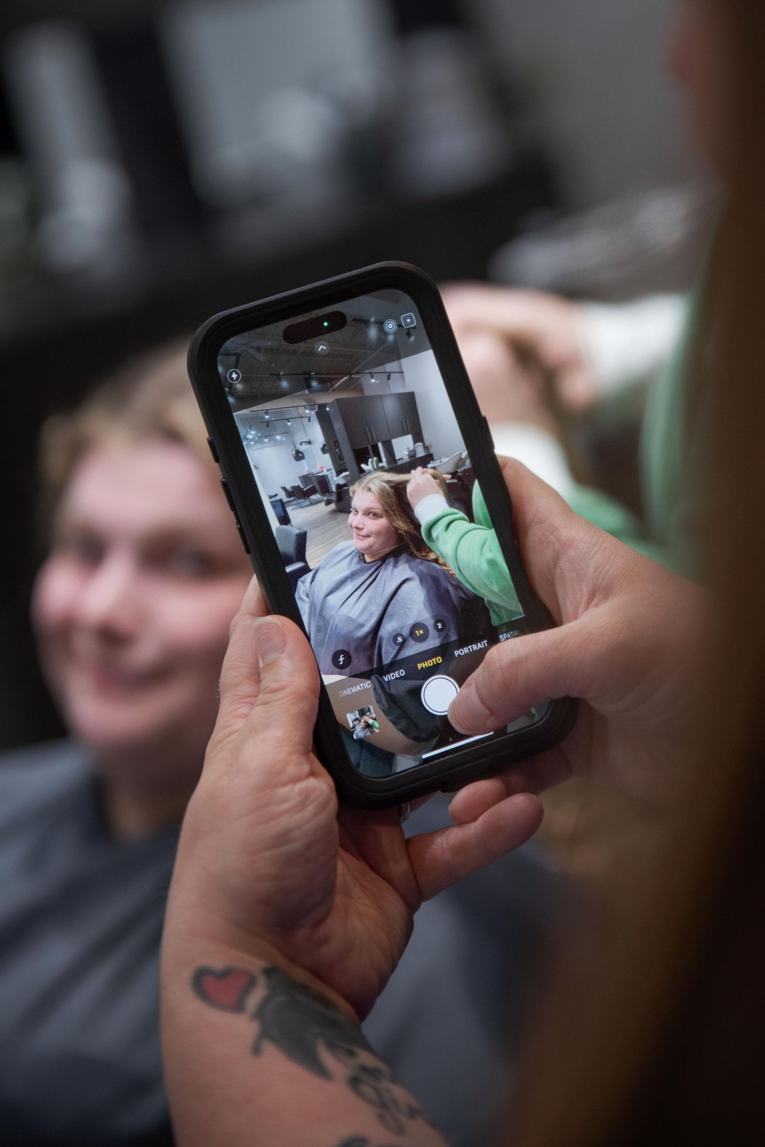 A tattooed hand holds a phone photographing a woman getting her hair styled in a salon.