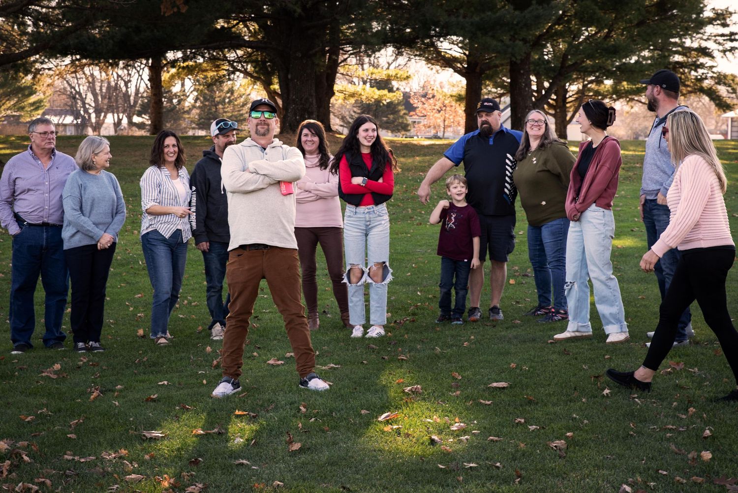 A large family group gathers outdoors on a green lawn surrounded by autumn trees for a photo session.