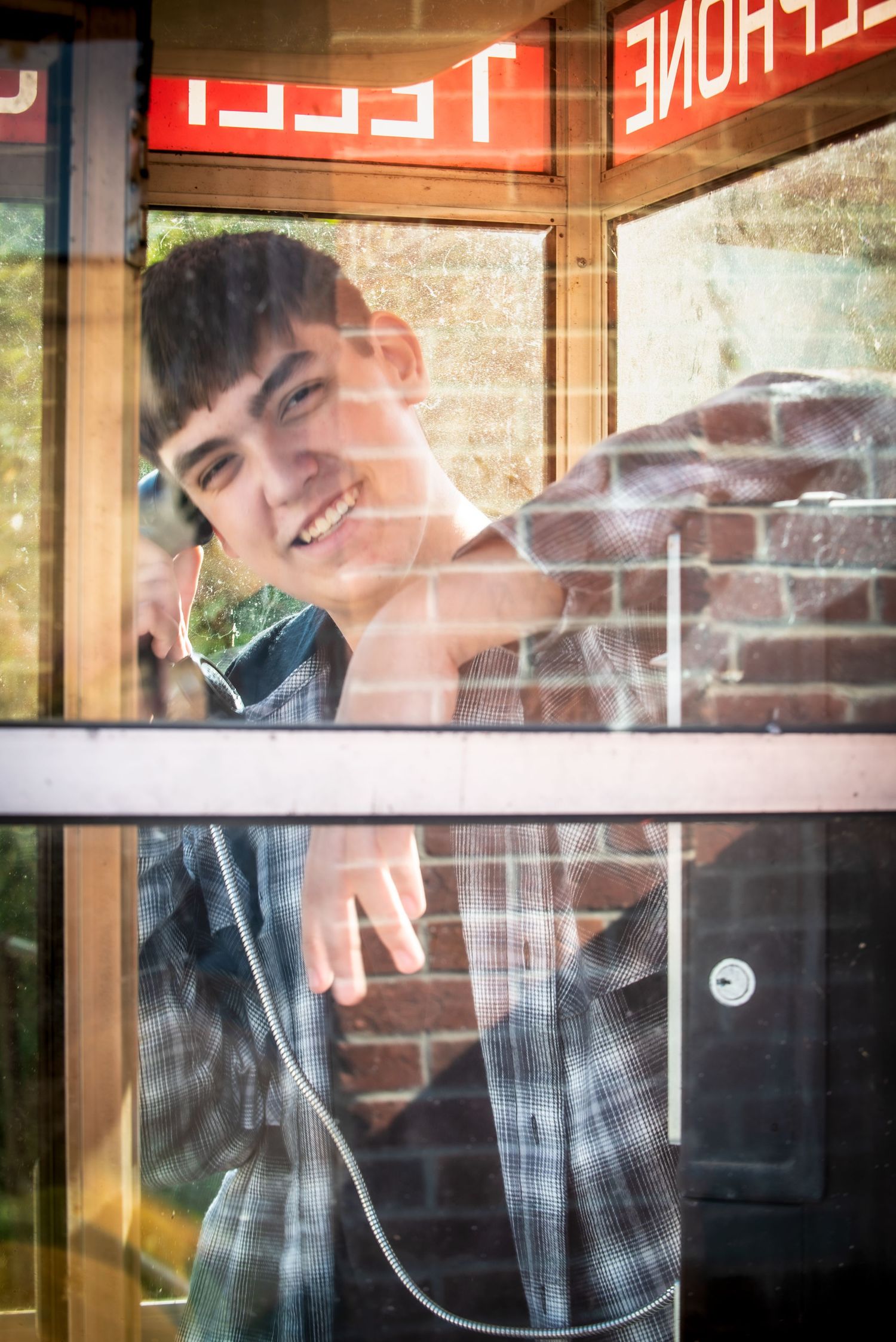 Smiling teenage boy inside a glass telephone booth reaching toward the camera.