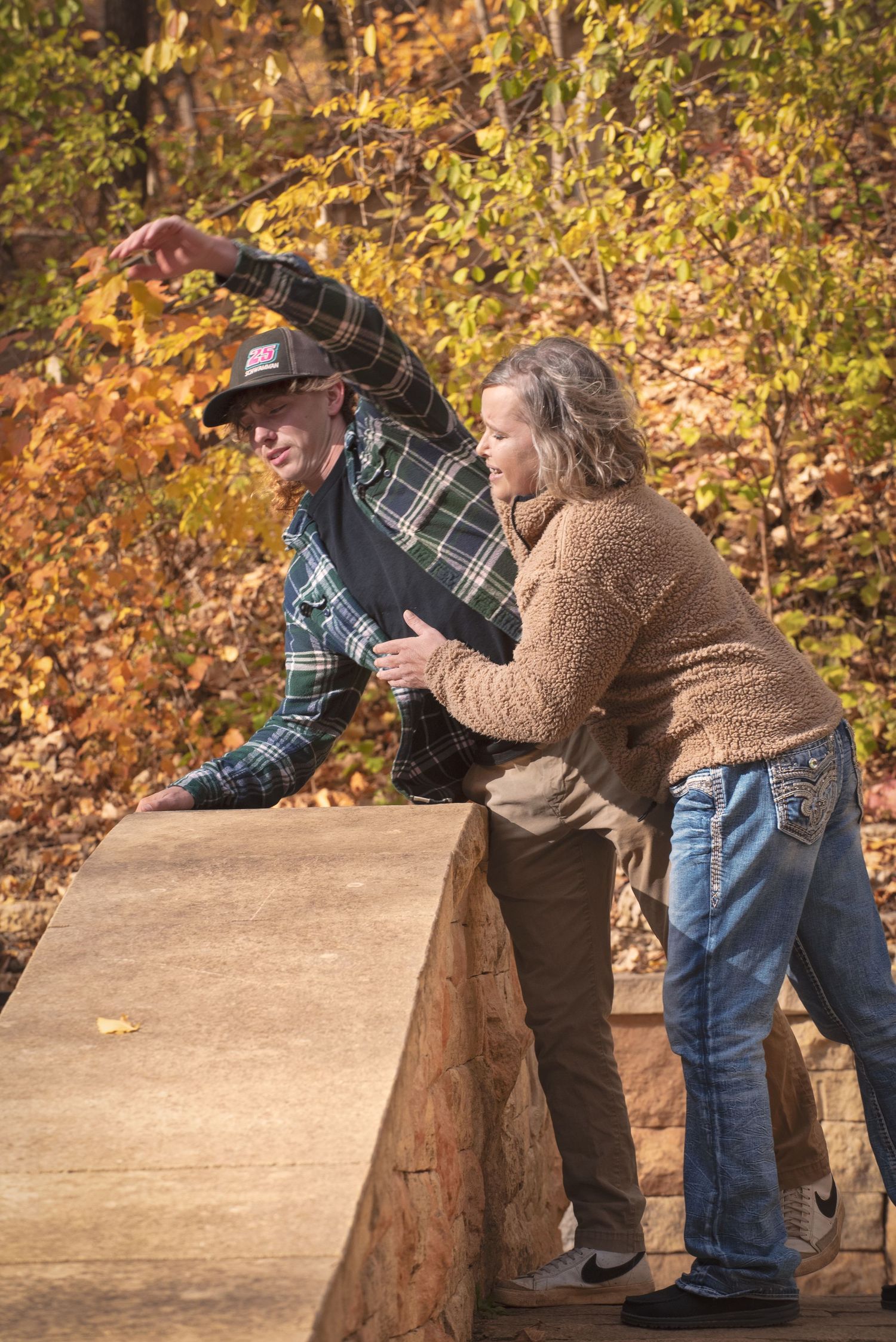 Couple leans over a stone bridge wall pointing at something in the distance amid fall foliage backdrop.