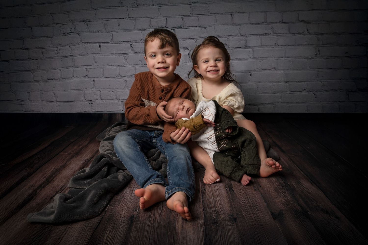 Two young siblings sitting on a wooden floor holding their newborn baby against a brick wall backdrop.