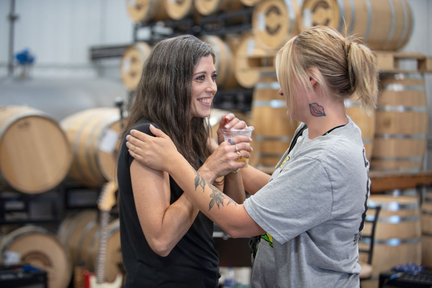 Two women smiling and sharing a drink together inside a distillery warehouse with wooden barrels at Harmony Spirits..