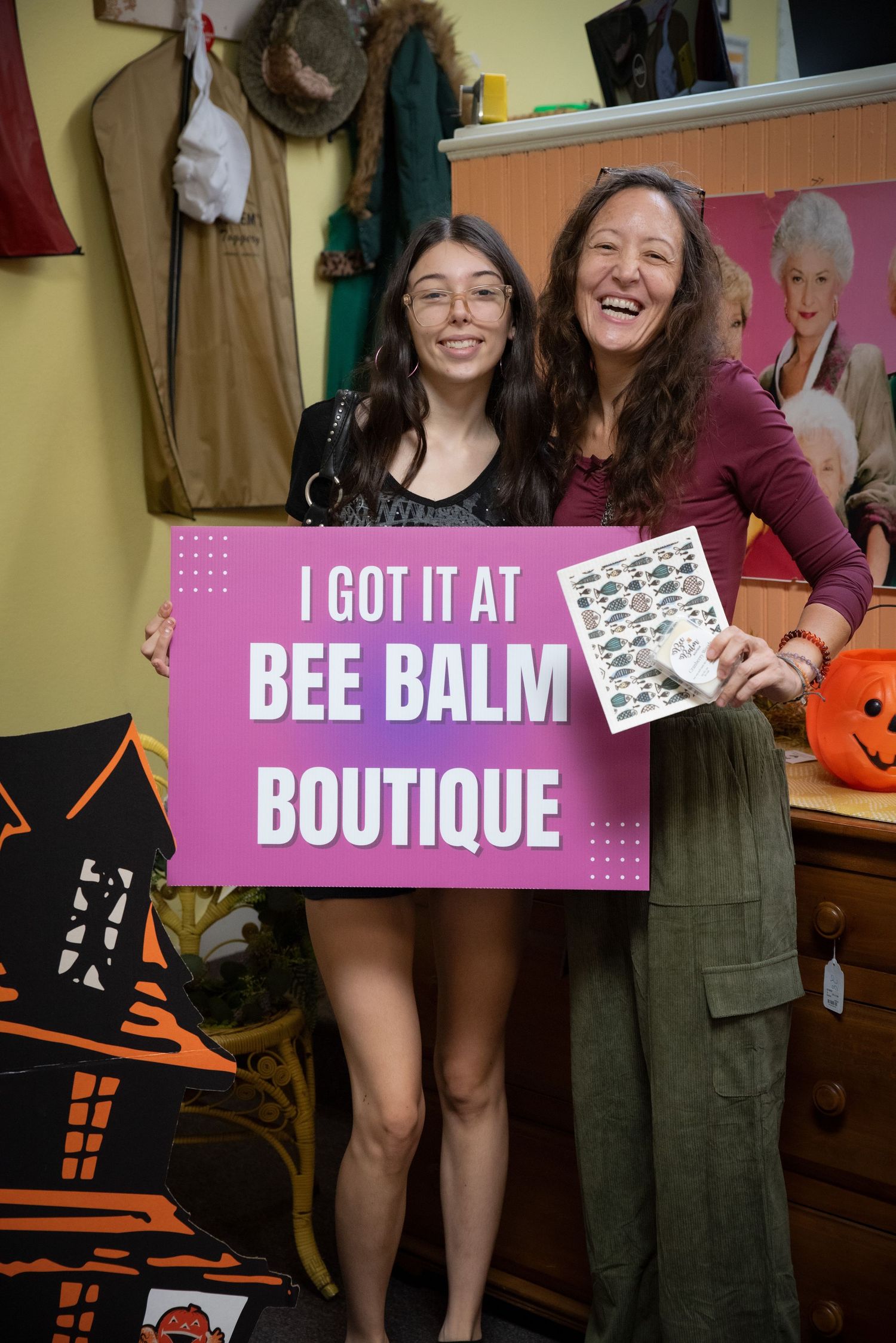 Two women smile holding a pink Bee Balm Boutique sign inside a colorful retail shop.