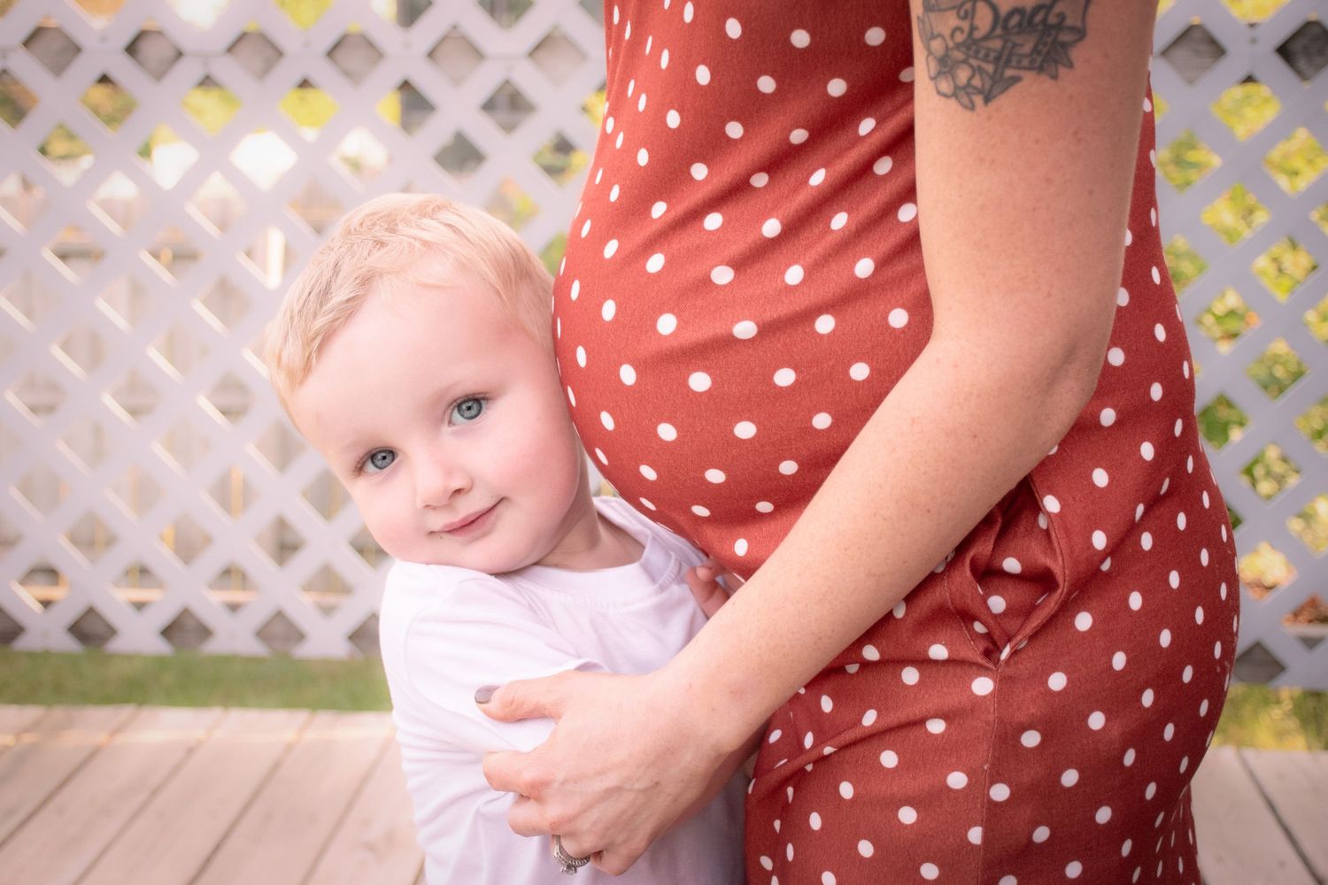 Adorable blonde toddler hugging pregnant mothers polka dot dress outdoors on wooden deck.