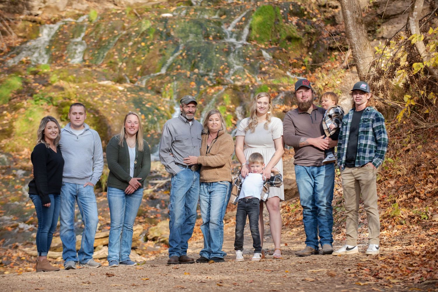 Family of eight poses along a rocky creek bed surrounded by colorful autumn trees and a small waterfall.