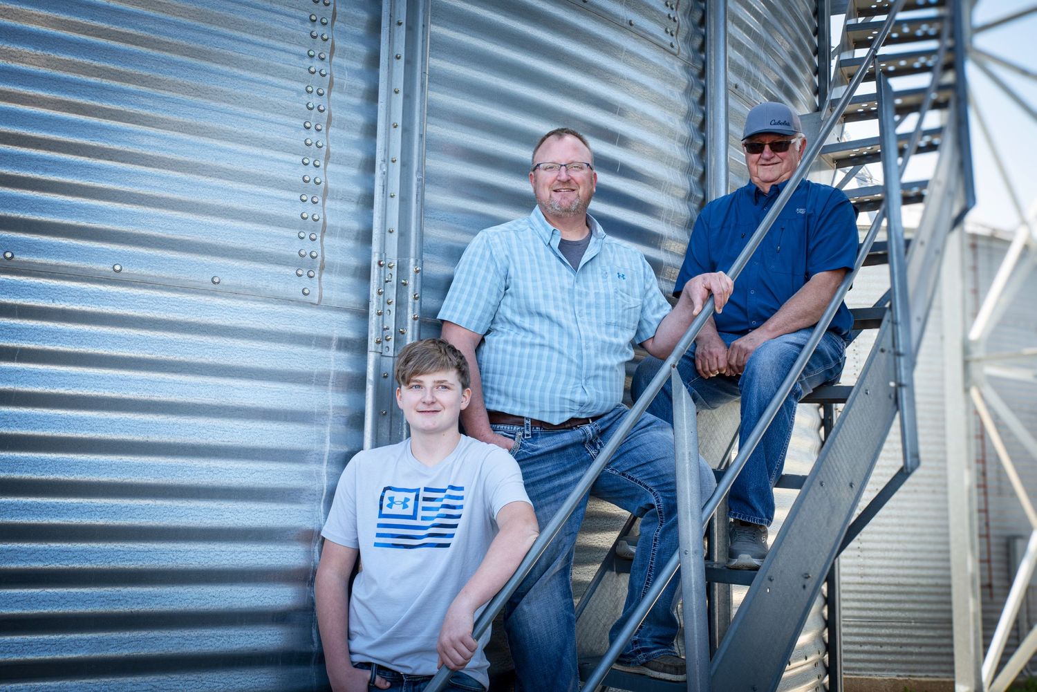 Three generations of men posing beside large metal grain storage bins on a farm.