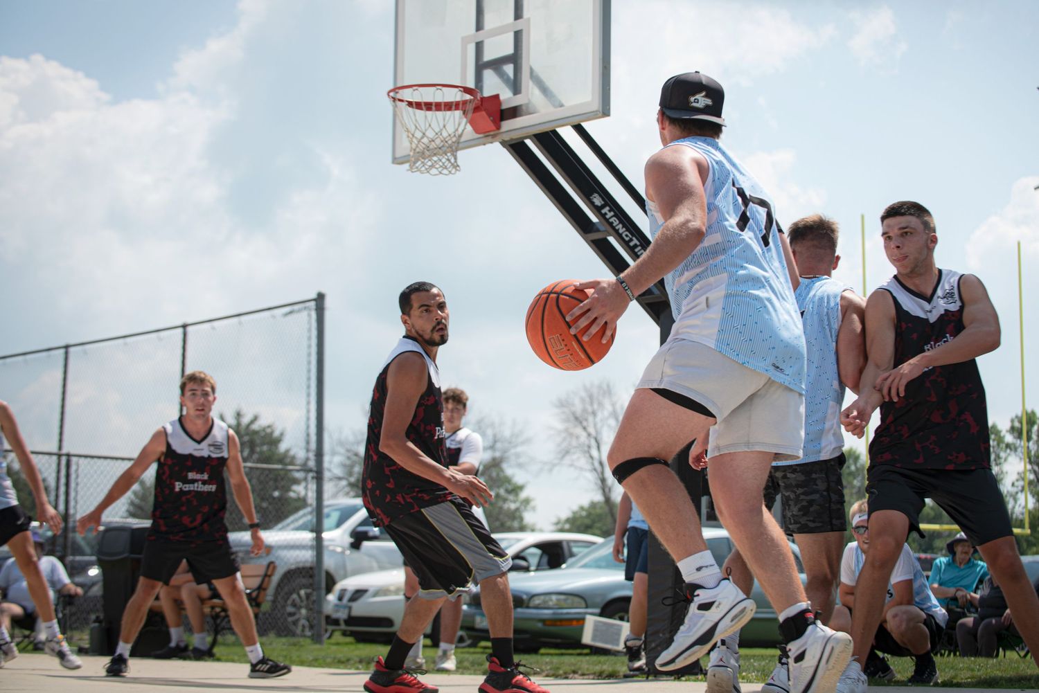 Young men play an intense outdoor basketball Rucker League game on a fenced court on a bright sunny day.