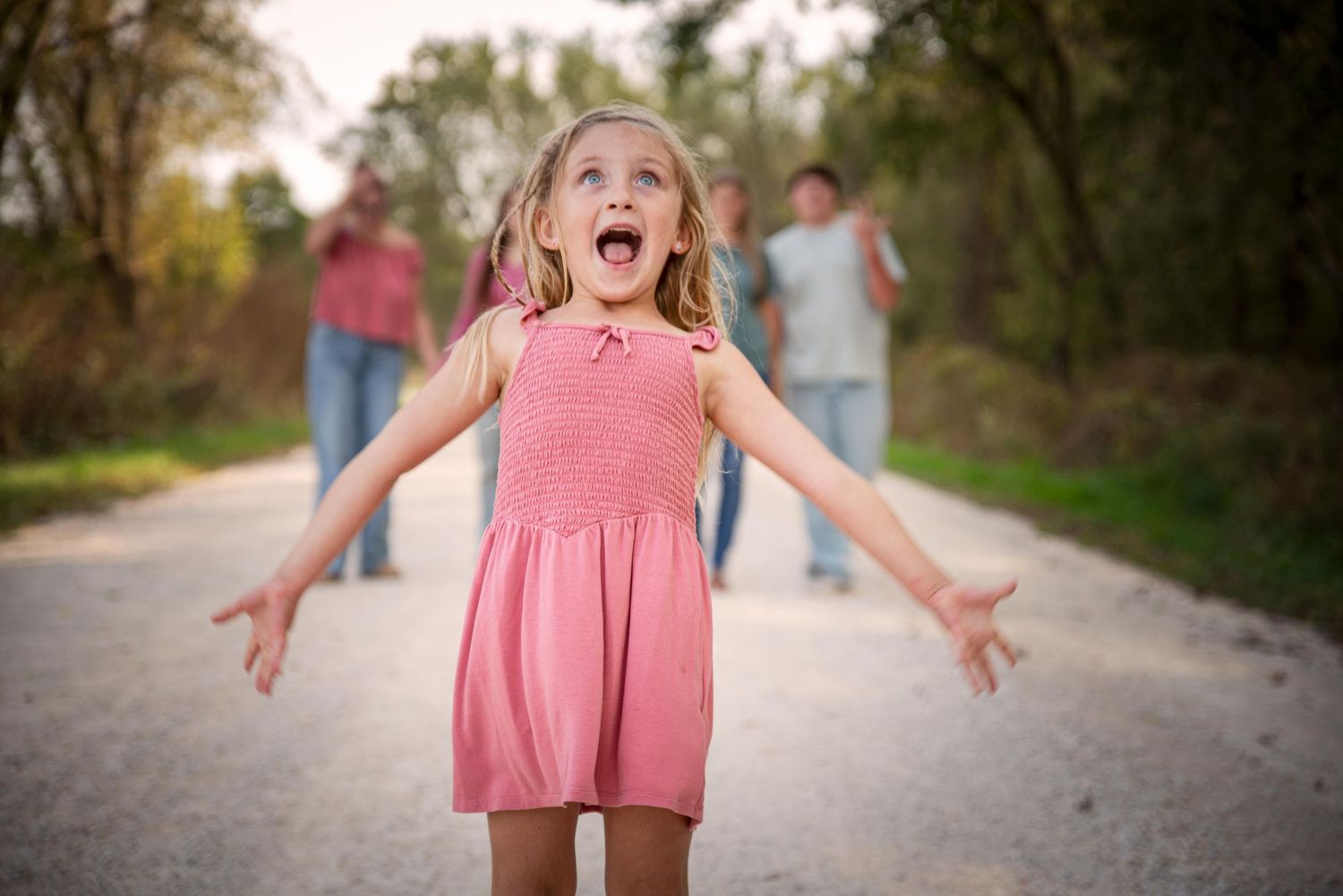 Excited young girl in pink dress with arms outstretched on a dirt path with family in background.