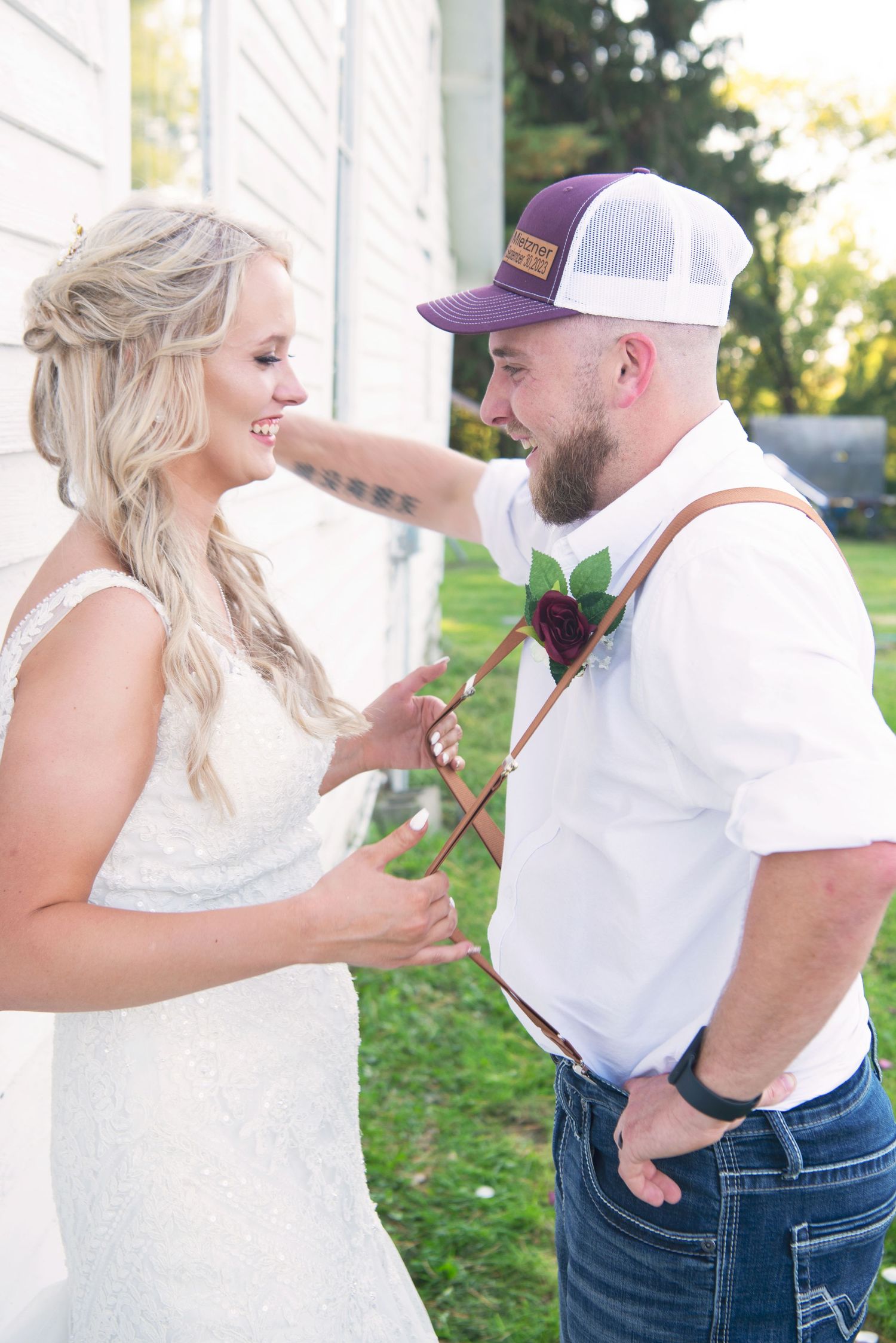 Bride in lace dress smiles as groom in suspenders and purple cap pins a boutonniere at rustic wedding.