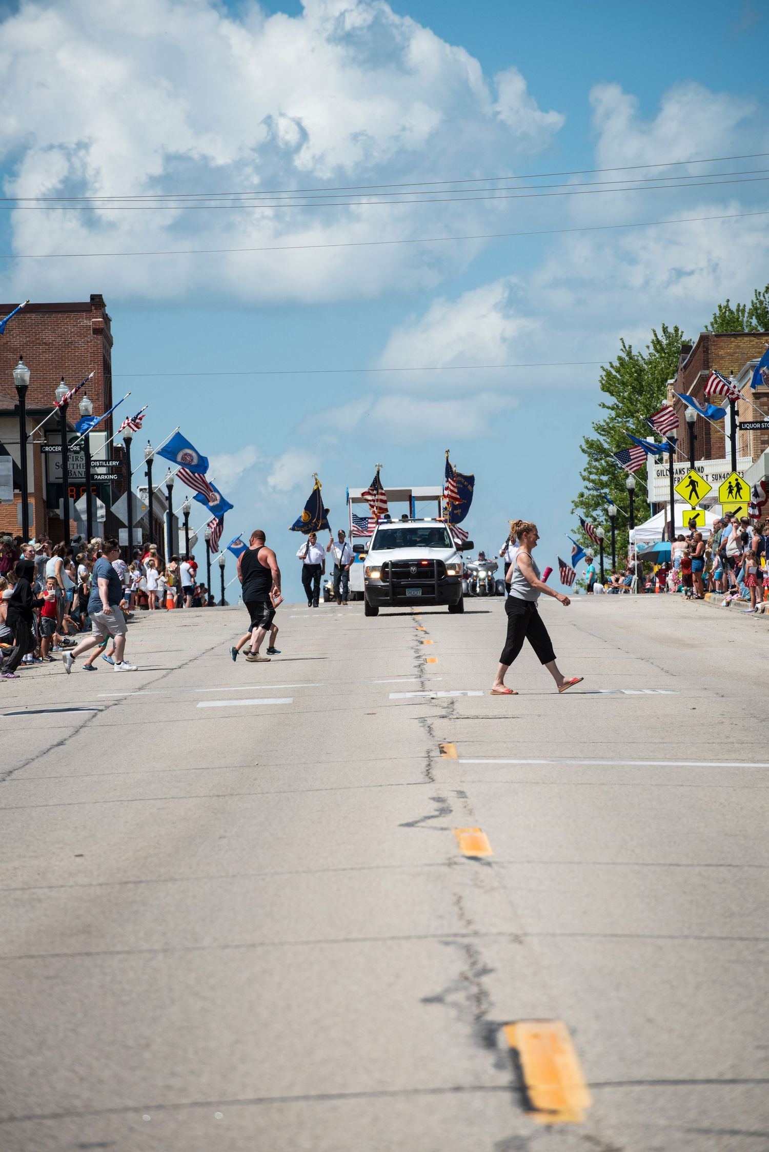 People cross a Main Street lined with American flags during a sunny small-town parade. At Harmony MN 4th of July Celebration.