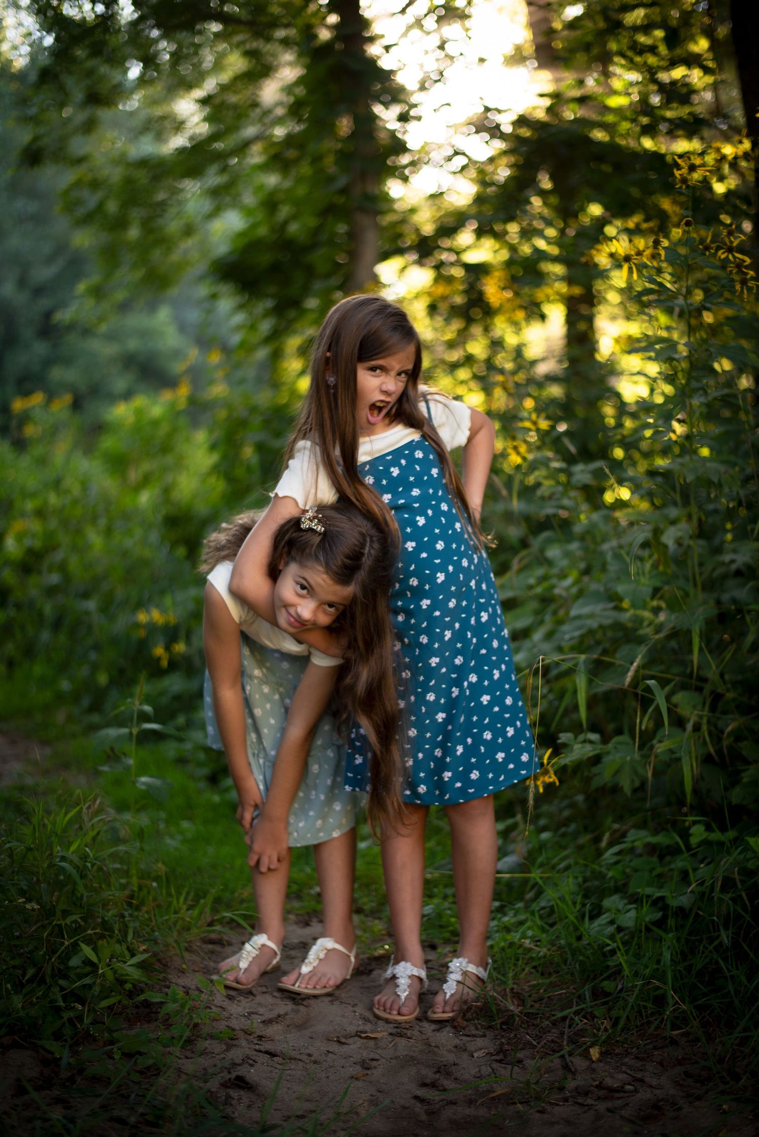 Two young girls playing in a lush green forest path, one piggyback riding while laughing outdoors.